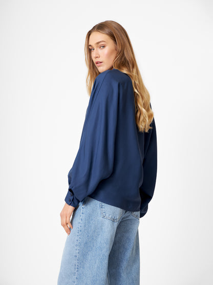 Eine Frau mit langen, gewellten blonden Haaren trägt die Bluse Antonia Slim Tencel Blue und hellblaue Jeans. Sie steht vor einem weißen Hintergrund und schaut über ihre Schulter.