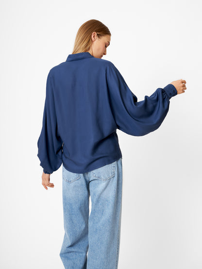 Eine Frau mit glattem blondem Haar blickt von sich weg. Sie trägt die Bluse Antonia Slim Tencel Blue und hellblaue Jeans vor einem schlichten weißen Hintergrund.