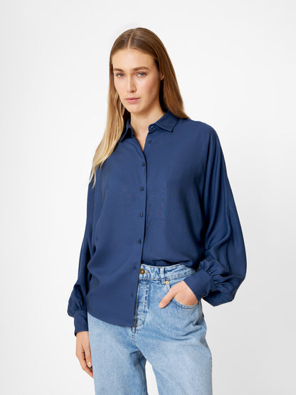 Eine Frau mit langen blonden Haaren trägt die Bluse Antonia Slim Tencel Blue und hellblaue Jeans. Sie steht mit einer Hand in der Hosentasche und blickt ruhig nach vorne auf einen schlichten weißen Hintergrund.