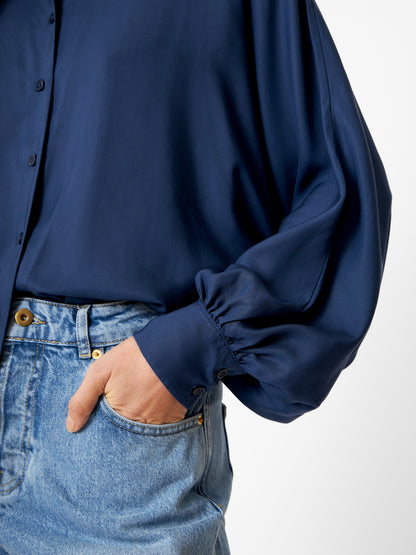 Eine Person trägt eine locker sitzende Bluse Antonia Slim Tencel Blue mit weiten Ärmeln, die in eine hellblaue High-Waisted-Jeans gesteckt ist, und steht mit einer Hand in der Tasche vor einem schlichten Hintergrund.