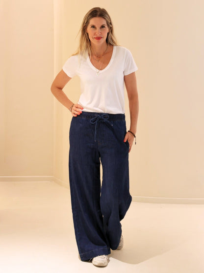 Eine Frau mit langen blonden Haaren in einem weißen T-Shirt, einer dunkelblauen Hose und weißen Turnschuhen steht in einem hellen, minimalistischen Raum und lächelt, eine Hand in der Tasche - perfekt für Liebhaber nachhaltiger Mode.