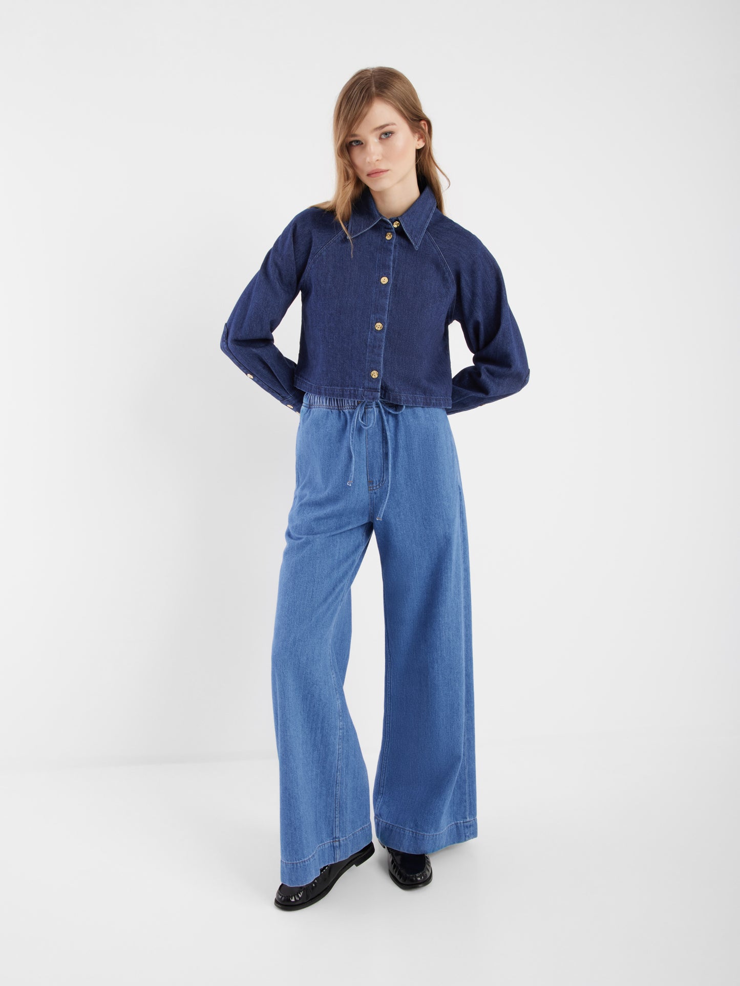 Eine Person steht vor einem unifarbenen weißen Hintergrund und trägt die dunkelblaue, langärmelige Jacke Hanna Denim und eine hellblaue Jeans mit hoher Taille und weitem Bein und schwarzen Schuhen - ein stilvoller Co-Ord-Look, perfekt für mühelosen Chic.