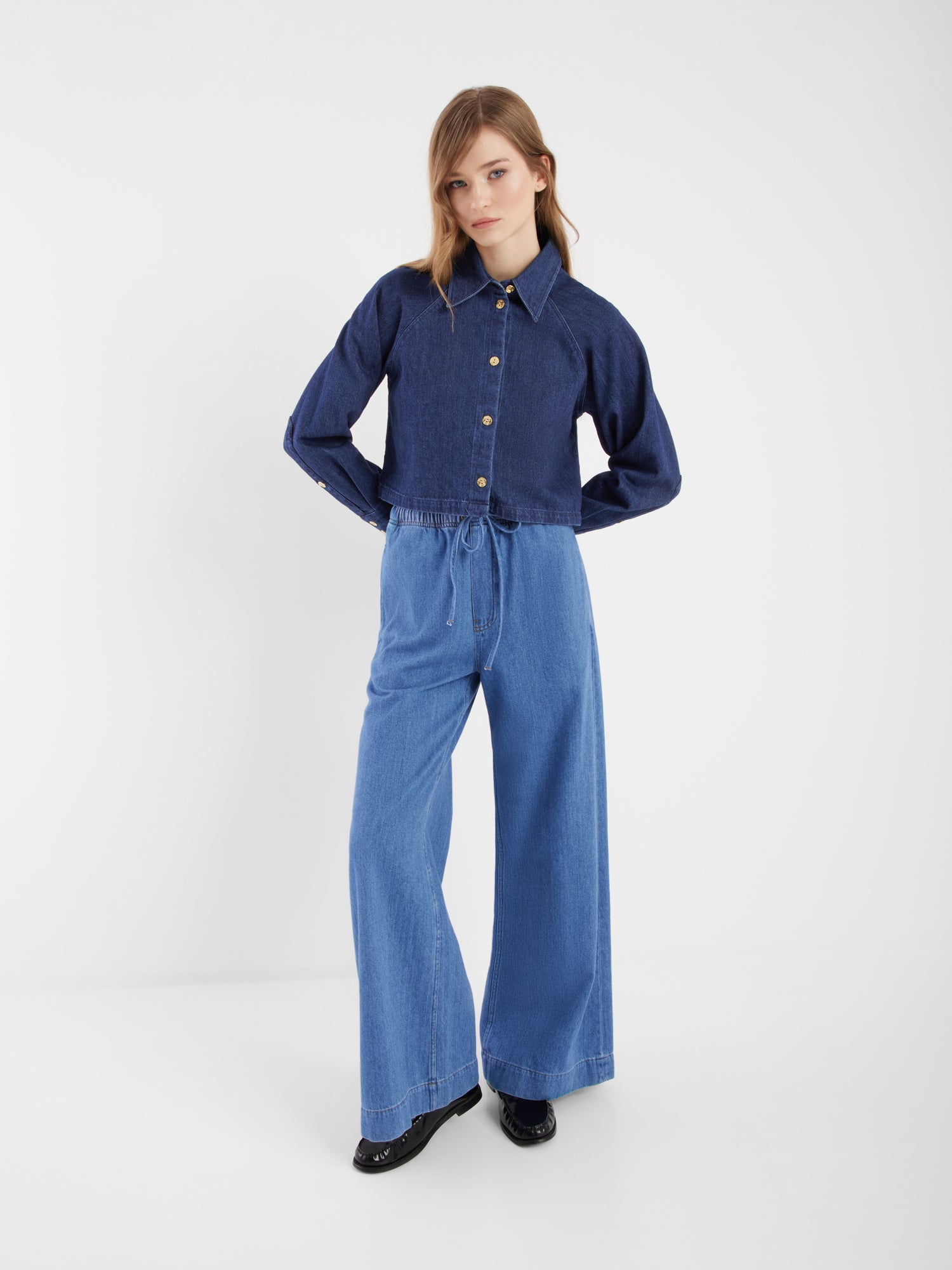 Eine Person steht vor einem unifarbenen weißen Hintergrund und trägt die dunkelblaue, langärmelige Jacke Hanna Denim und eine hellblaue Jeans mit hoher Taille und weitem Bein und schwarzen Schuhen - ein stilvoller Co-Ord-Look, perfekt für mühelosen Chic.