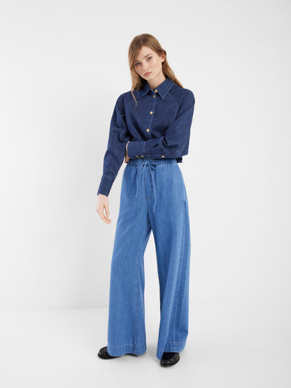 Eine Frau posiert lässig vor einem schlichten weißen Hintergrund in der Jacke Hanna Denim, die in eine hellblaue Jeans mit hoher Taille und weitem Bein gesteckt und mit schwarzen Schuhen kombiniert wird - und zeigt mit ihrem langen hellbraunen Haar einen entspannten Co-Ord Look.