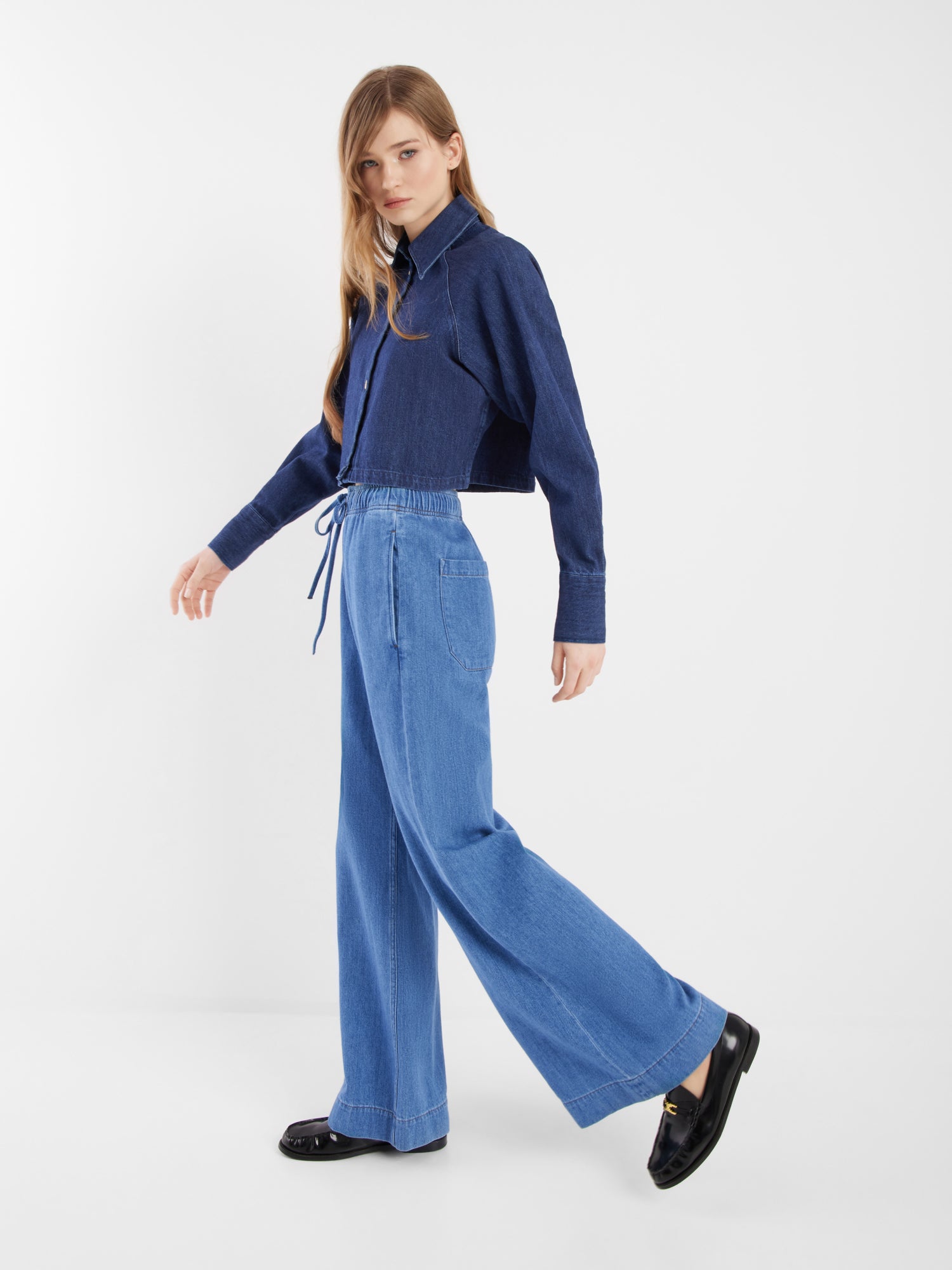 Eine Frau mit langem, glattem Haar trägt die dunkelblaue Jacke Hanna Denim und hellblaue Jeans mit weitem Bein für einen schicken Kombinationslook mit schwarzen Halbschuhen vor einem schlichten weißen Hintergrund.