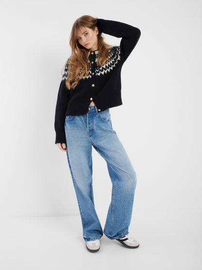 Eine Frau posiert lässig mit der Jacke Norweger Vol II Darkblue, hellblauen Jeans mit weitem Bein und weißen Turnschuhen vor einem schlichten weißen Hintergrund.