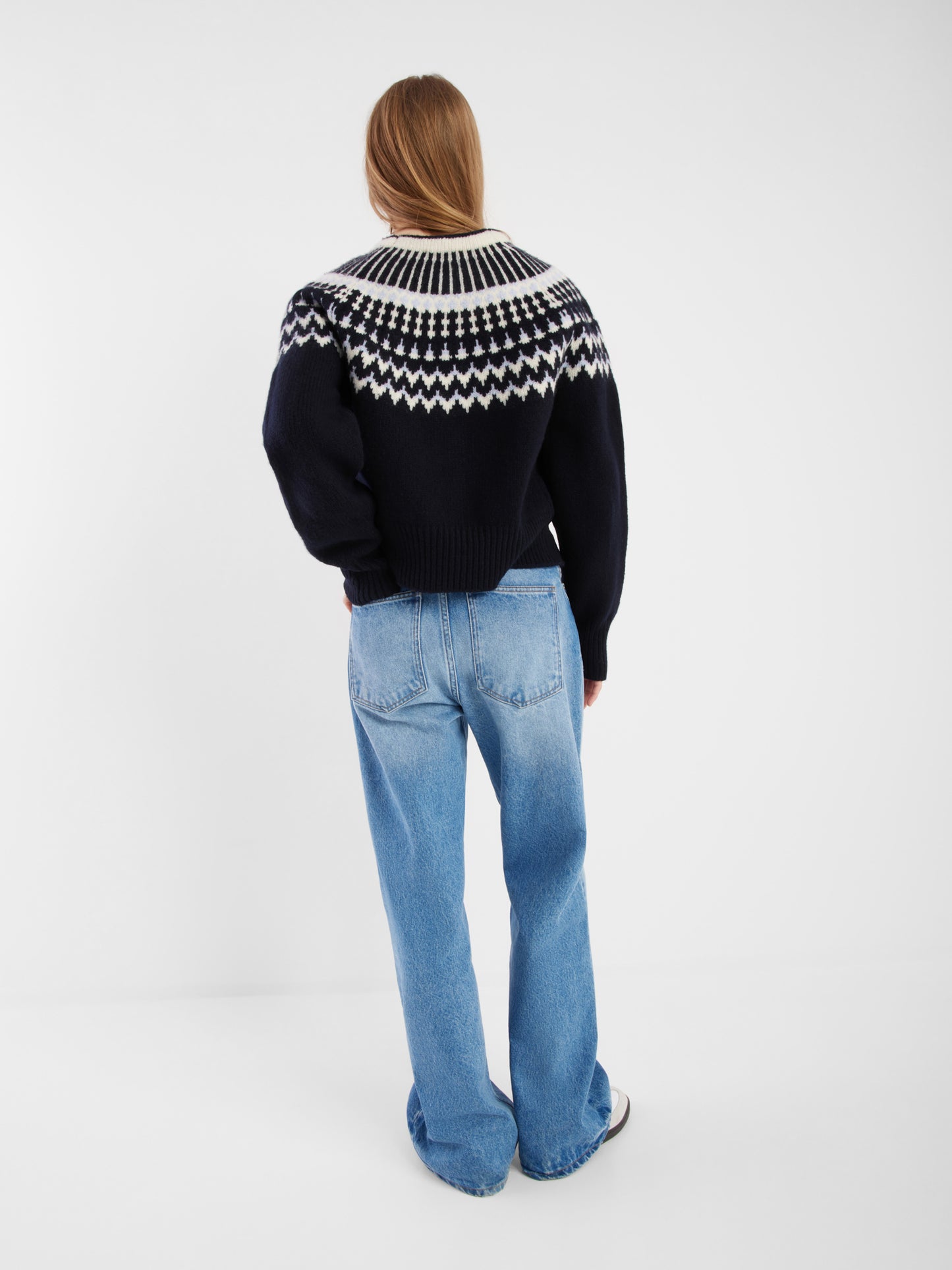 Eine Person steht mit dem Rücken zur Kamera und trägt die Jacke Norweger Vol II Darkblue, hellblaue Jeans und Turnschuhe. Ihr langes hellbraunes Haar ist vor einem schlichten weißen Hintergrund zu sehen.