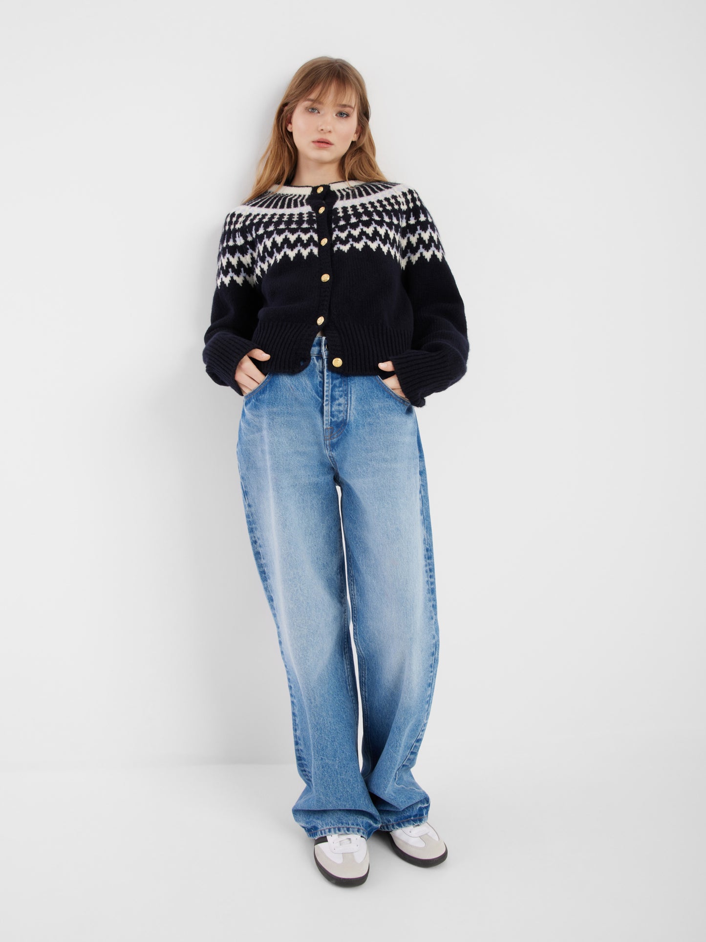 Eine junge Frau steht vor einem schlichten weißen Hintergrund und trägt die Jacke Norweger Vol II Darkblue, hellblaue Jeans mit weitem Bein und weiße Turnschuhe, die Hände in den Taschen.