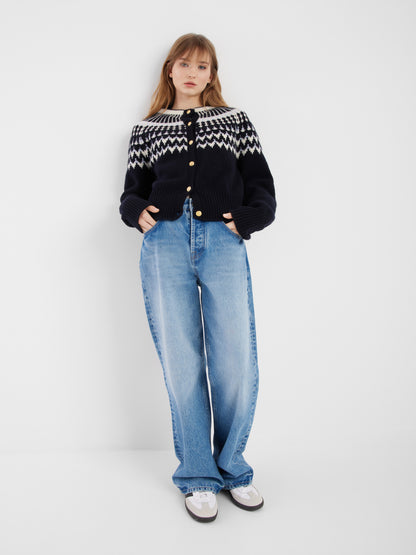 Eine junge Frau steht vor einem schlichten weißen Hintergrund und trägt die Jacke Norweger Vol II Darkblue, hellblaue Jeans mit weitem Bein und weiße Turnschuhe, die Hände in den Taschen.