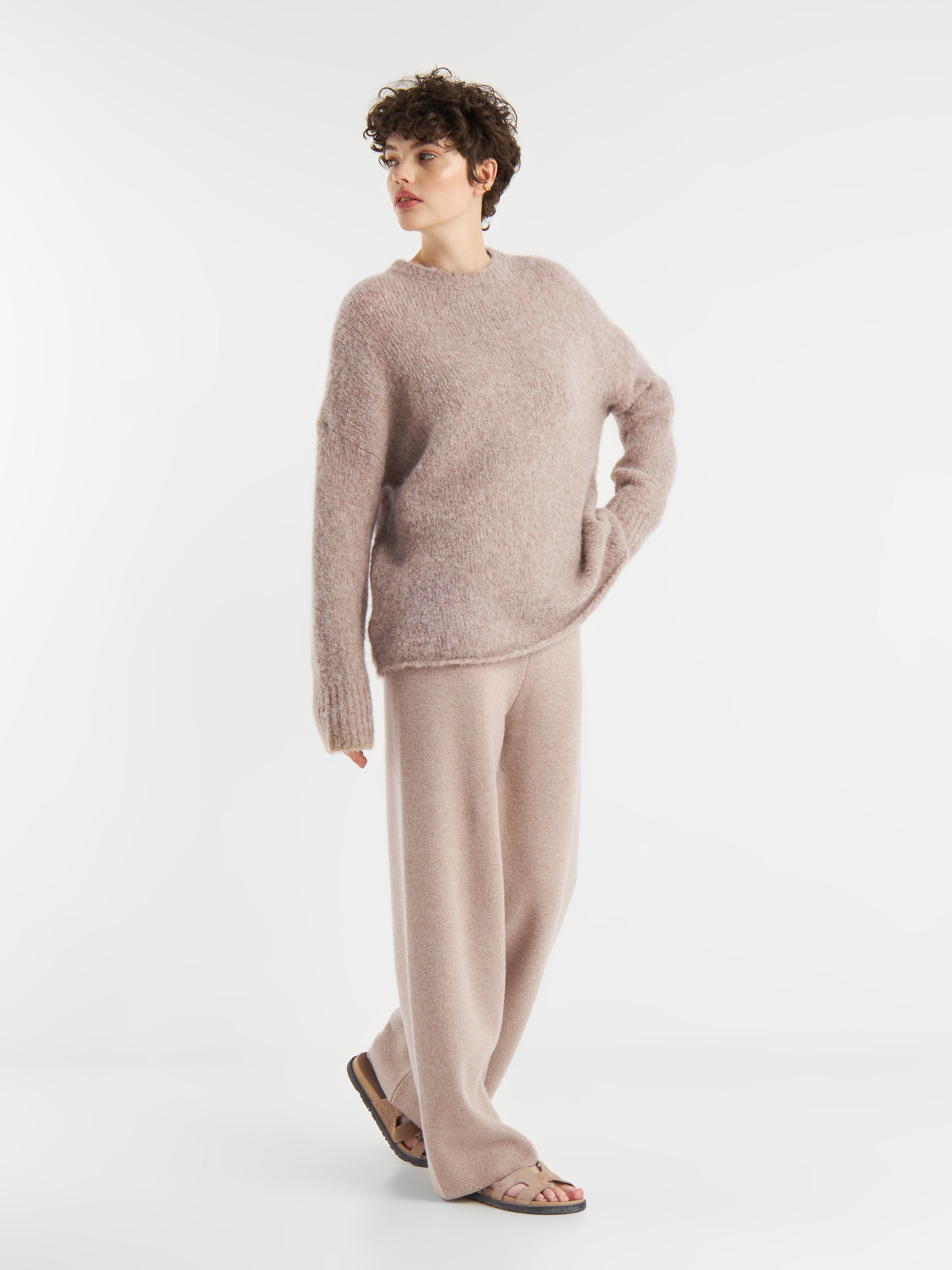 Eine Person mit kurzen lockigen Haaren trägt den Pullover LIV Beige und eine dazu passende Hose mit weitem Bein und Sandalen. Sie steht vor einem schlichten weißen Hintergrund und schaut zur Seite.
