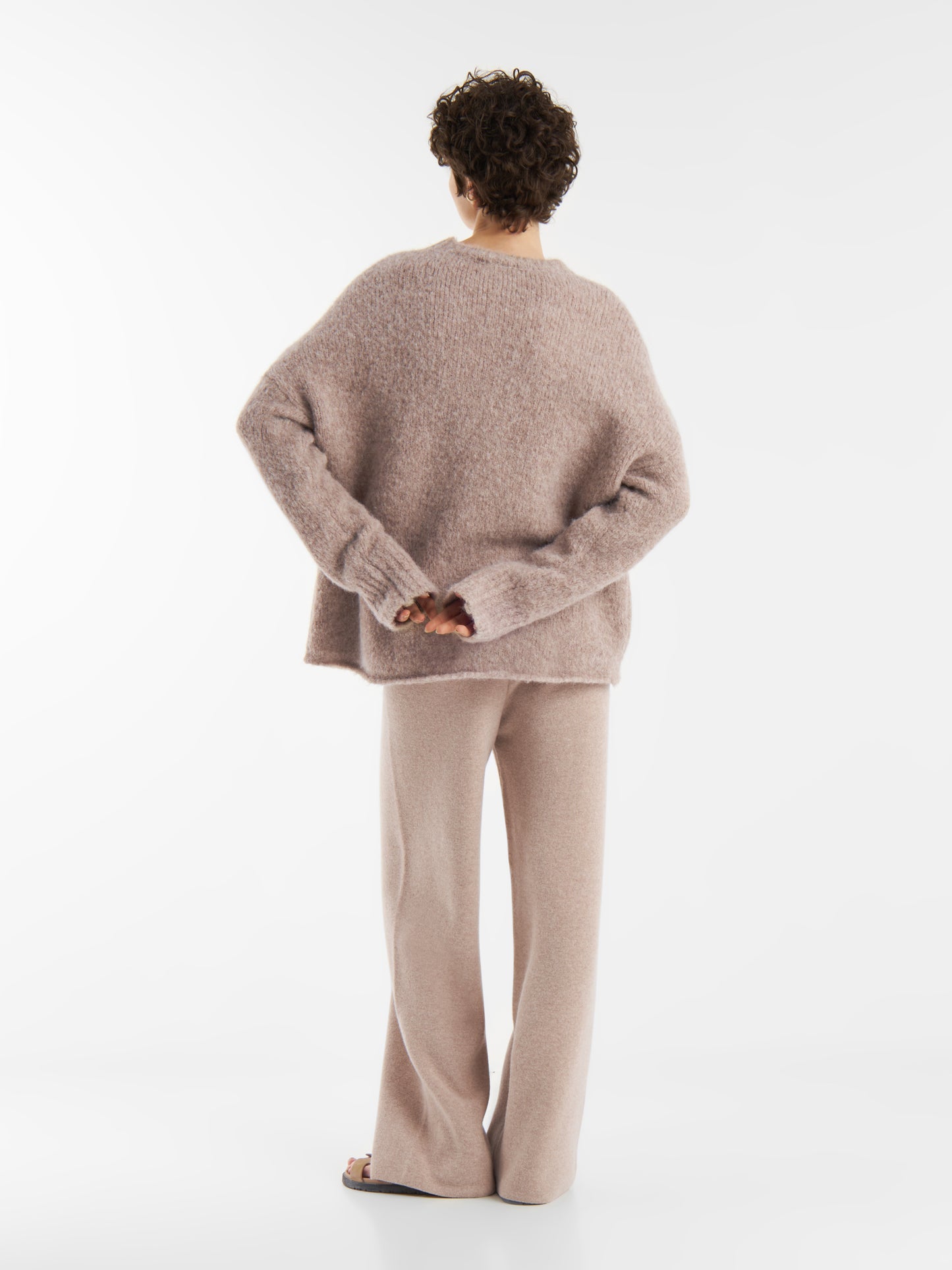 Eine Person mit kurzen lockigen Haaren, von hinten gesehen, trägt den lockeren, weich strukturierten Pullover LIV Beige mit einer passenden Hose mit weitem Bein. Ihre Hände sind hinter dem Rücken verschränkt und stehen vor einem schlichten weißen Hintergrund.