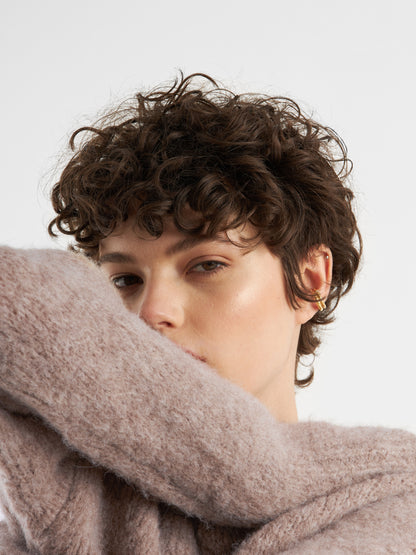 Eine Person mit kurzen, lockigen braunen Haaren und goldenen Ohrringen blickt in die Kamera und verdeckt ihr Gesicht teilweise mit einem hochgezogenen, flauschigen Pullover LIV Beige vor einem schlichten weißen Hintergrund.