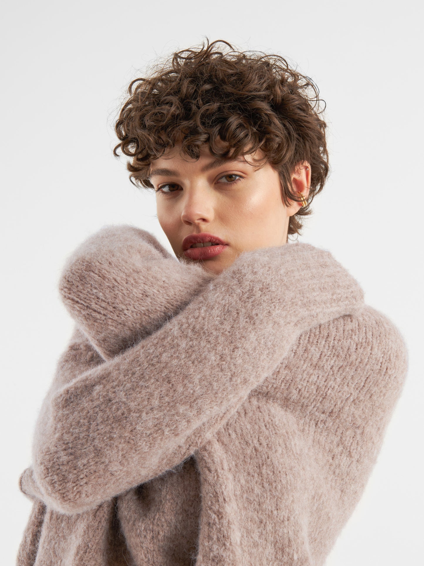 Eine Person mit kurzen, lockigen braunen Haaren trägt den Pullover Liv Turtle Beige, verschränkt die Arme und schaut direkt in die Kamera vor einem schlichten weißen Hintergrund.