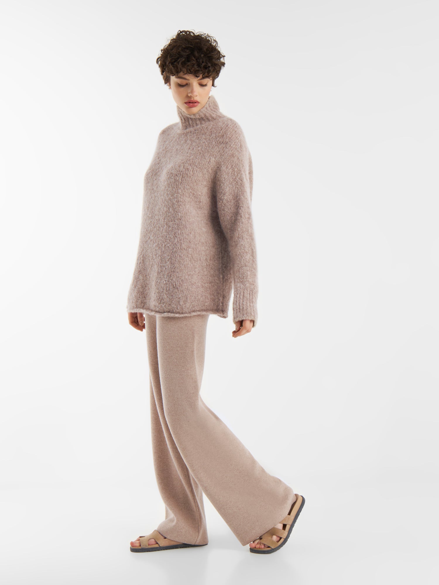 Eine Person, die den Pullover LIV Beige trägt, locker und kuschelig mit einem Rollkragen, gestylt mit einer passenden Hose mit weitem Bein und beigen Sandalen, steht vor einem schlichten weißen Hintergrund und blickt über ihre Schulter.