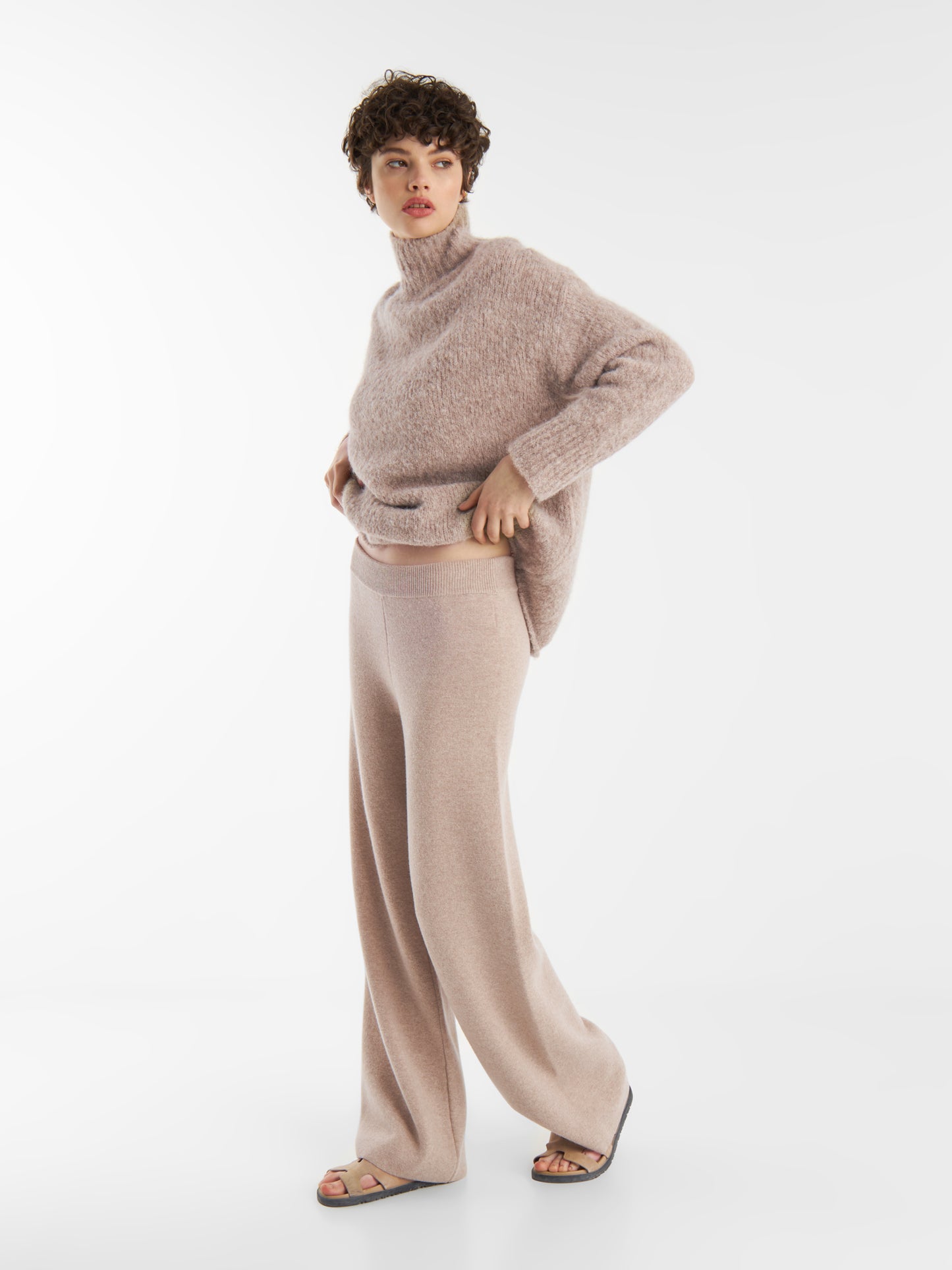 Eine Person, die den Pullover LIV Beige und eine Hose mit weitem Bein trägt, steht vor einem schlichten weißen Hintergrund. Sie trägt außerdem Sandalen und schaut zur Seite.