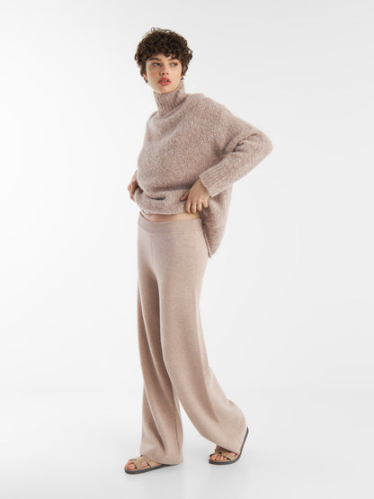 Eine Person, die den Pullover LIV Beige und eine Hose mit weitem Bein trägt, steht vor einem schlichten weißen Hintergrund. Sie trägt außerdem Sandalen und schaut zur Seite.