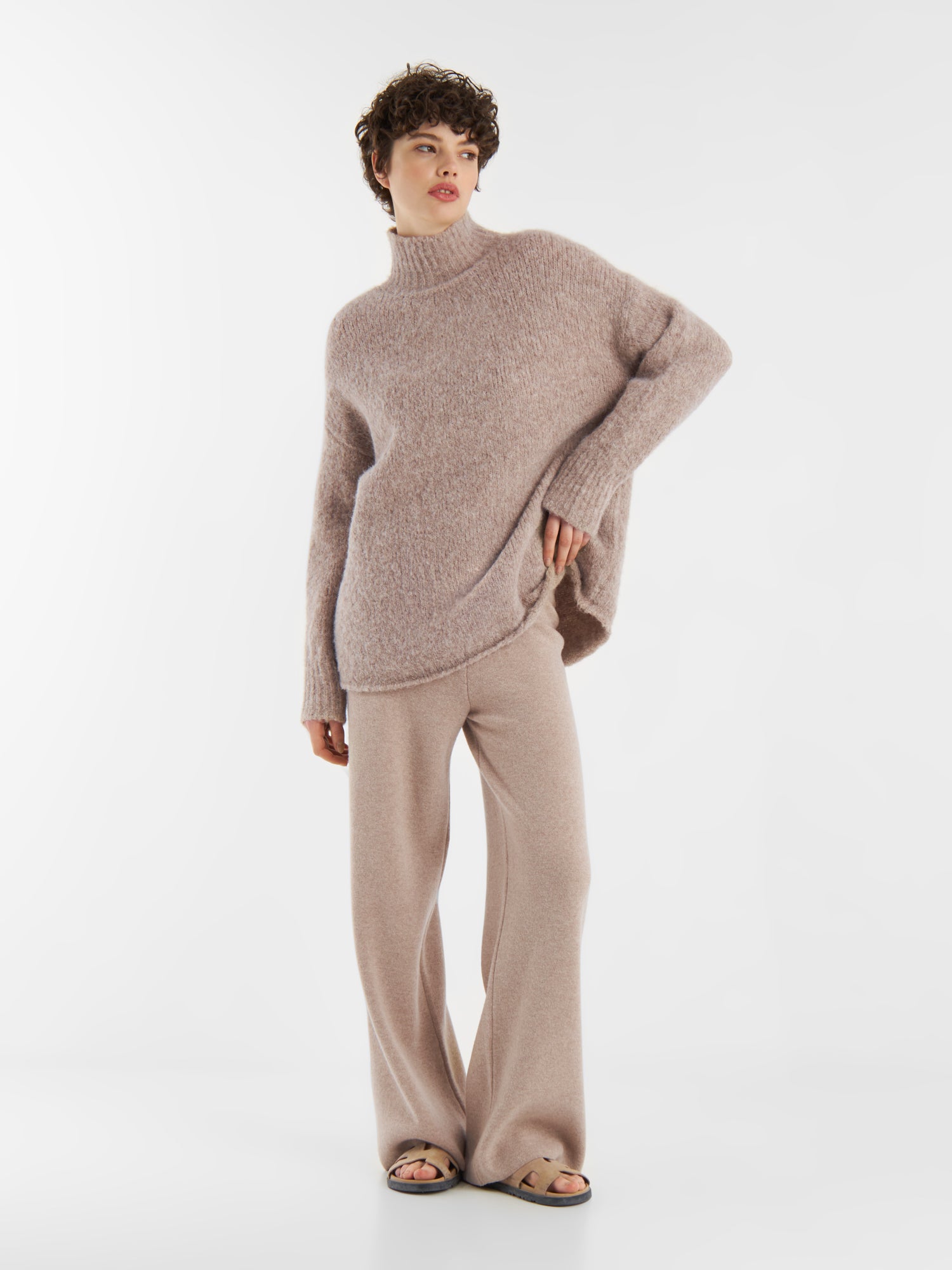 Eine Person steht vor einem einfarbig weißen Hintergrund und trägt den Pullover Liv Turtle Beige, eine passende Strickhose mit weitem Bein und Sandalen mit offenen Zehen.