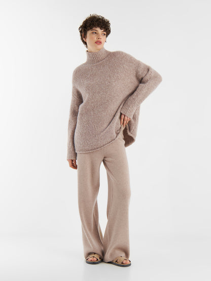 Eine Person steht vor einem einfarbig weißen Hintergrund und trägt den Pullover Liv Turtle Beige, eine passende Strickhose mit weitem Bein und Sandalen mit offenen Zehen.