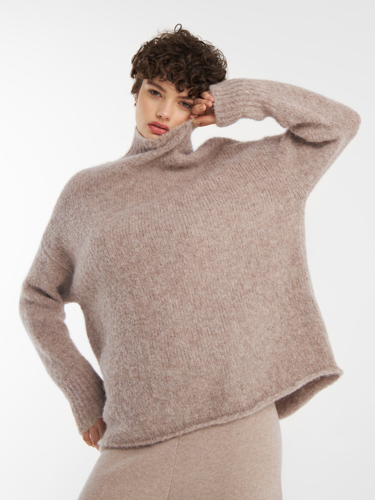 Eine Person mit kurzen lockigen Haaren posiert vor einem schlichten weißen Hintergrund, trägt den lockeren, weichen Pullover Liv Turtle Beige und den dazu passenden Rock und hat eine Hand auf die Stirn gelegt.