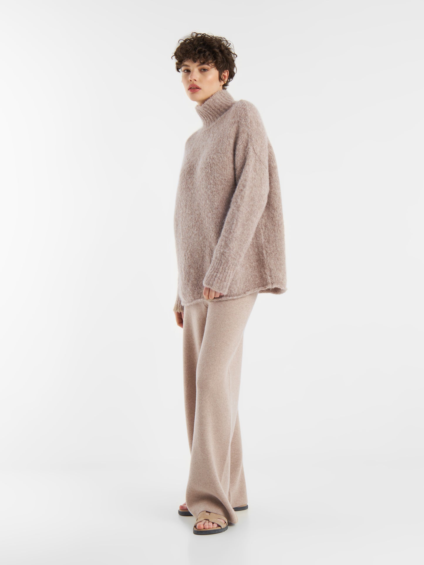 Eine Person mit kurzen lockigen Haaren trägt den Pullover Liv Turtle Beige, eine beigefarbene Hose mit weitem Bein und Sandalen mit offenen Zehen, die vor einem schlichten weißen Hintergrund stehen.