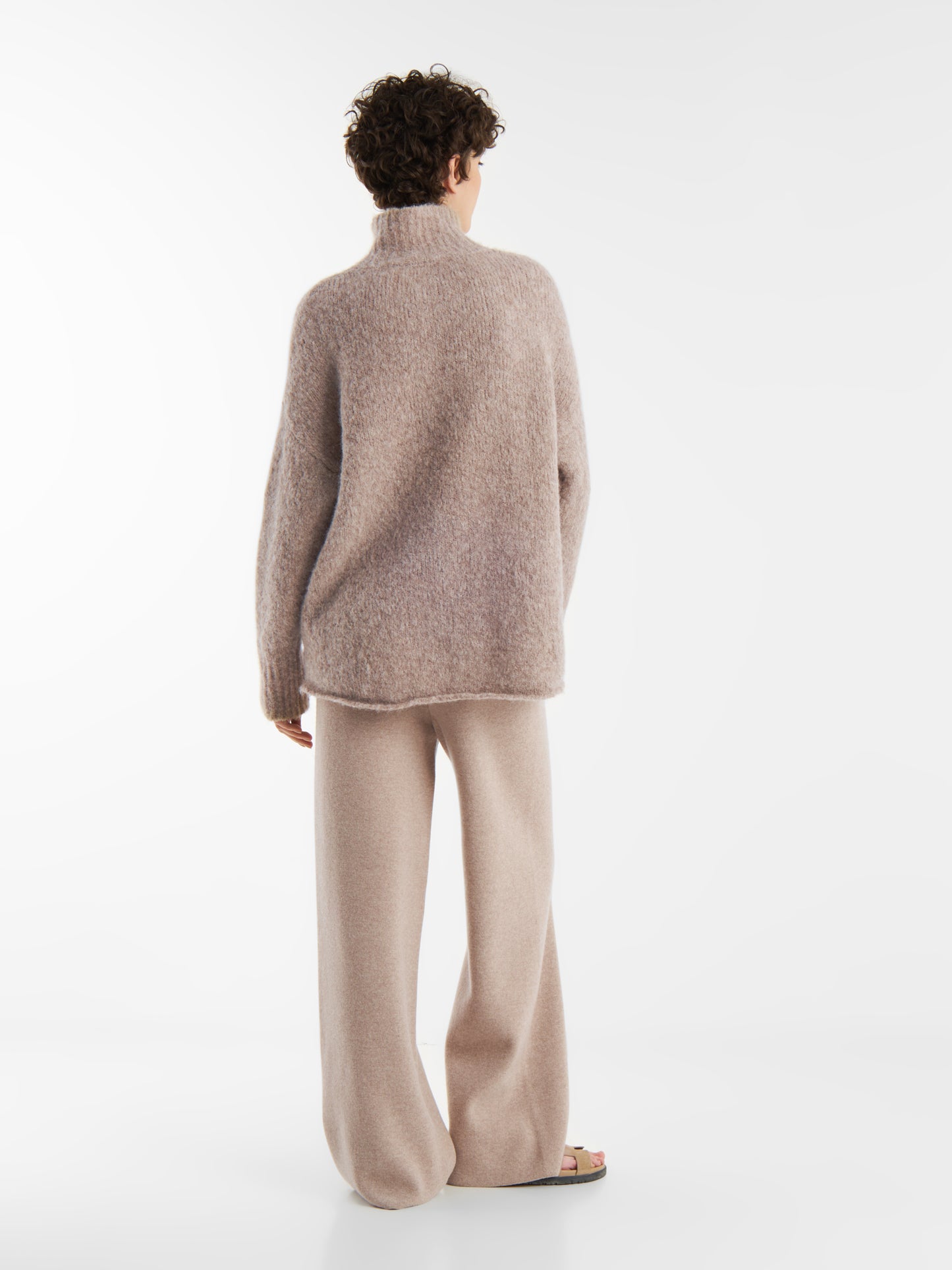 Eine Person mit kurzen lockigen Haaren steht mit dem Rücken zur Wand und trägt den Pullover Liv Turtle Beige sowie eine passende Hose mit weitem Bein und Sandalen. Der Hintergrund ist einfarbig weiß.