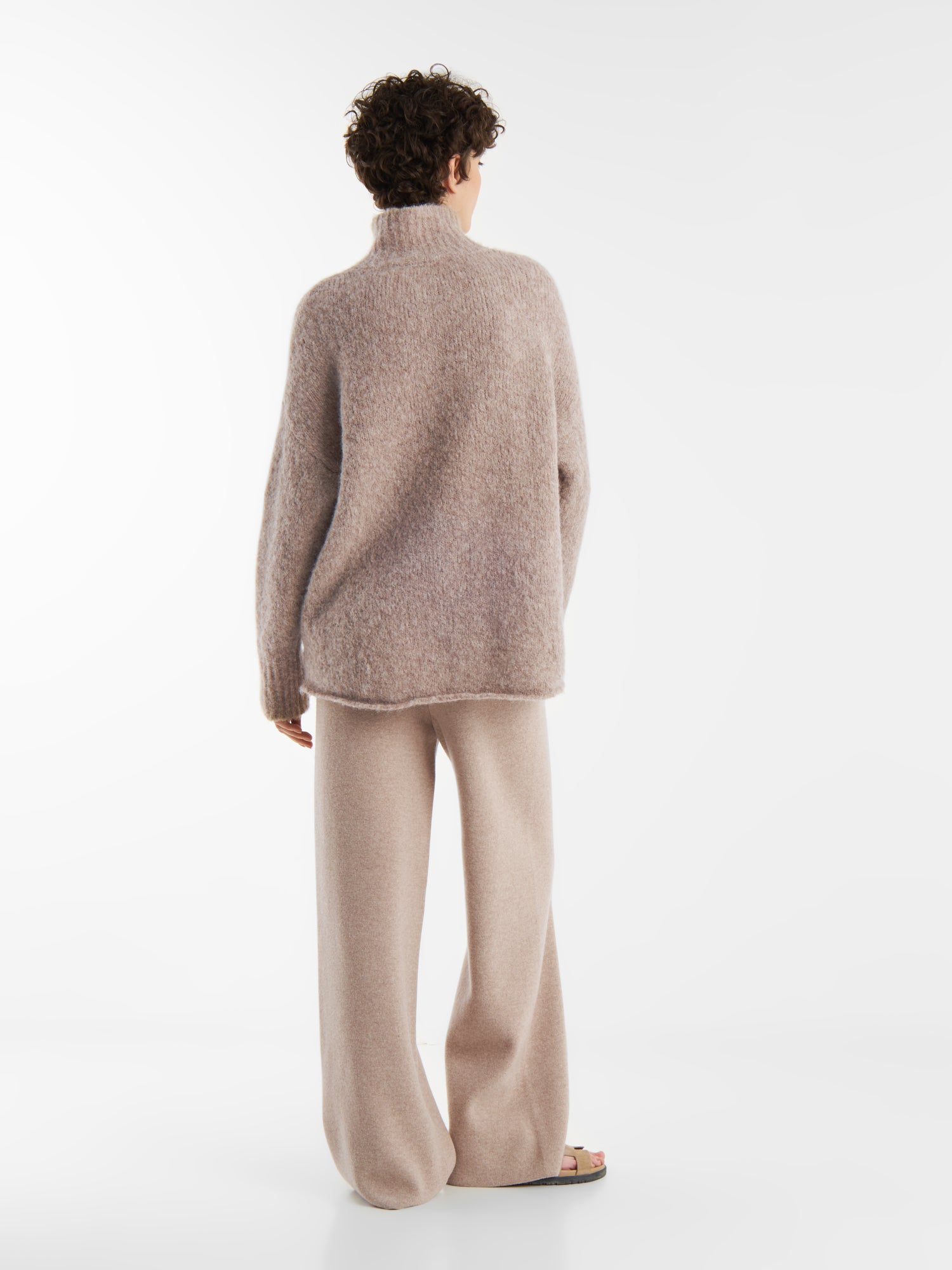 Eine Person mit kurzen lockigen Haaren steht mit dem Rücken zur Wand und trägt den Pullover Liv Turtle Beige sowie eine passende Hose mit weitem Bein und Sandalen. Der Hintergrund ist einfarbig weiß.