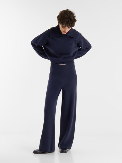 Eine Person mit kurzen lockigen Haaren steht leicht abgewandt und trägt ein dunkelblaues, langärmeliges Oberteil mit breitem Kragen und eine dazu passende dunkelblaue Merino-Hose vor einem schlichten weißen Hintergrund.