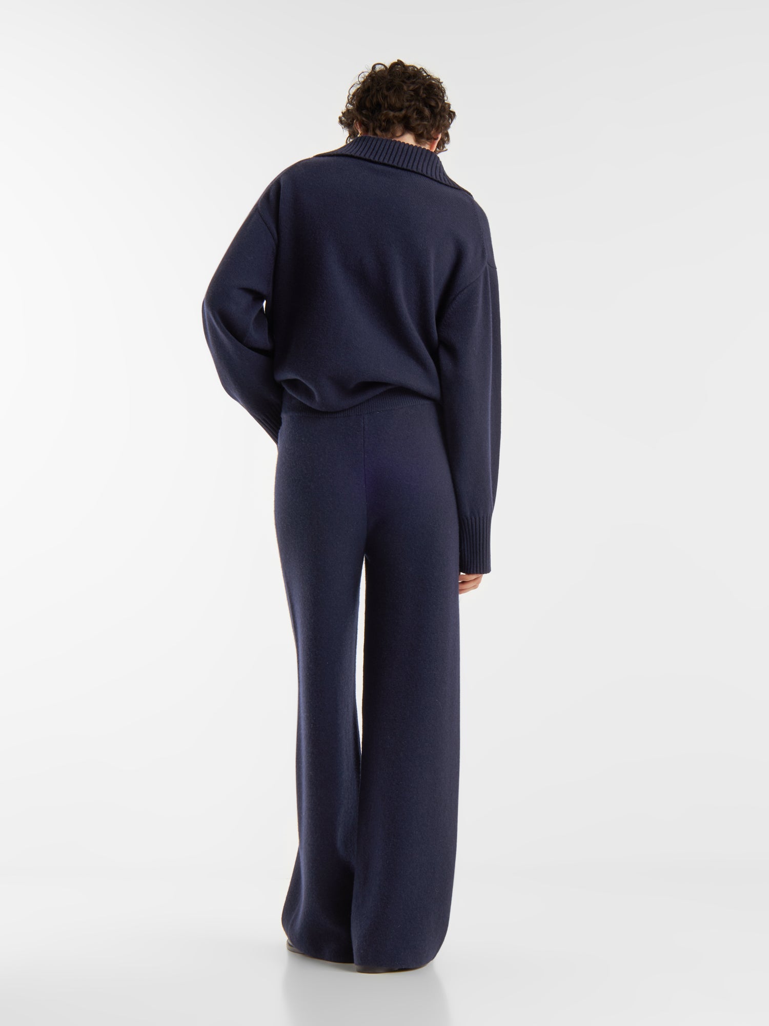 Eine Person steht mit dem Gesicht zur Kamera, trägt einen dunkelblauen Pullover Polo Merino und eine passende Hose mit weitem Bein vor einem schlichten weißen Hintergrund.