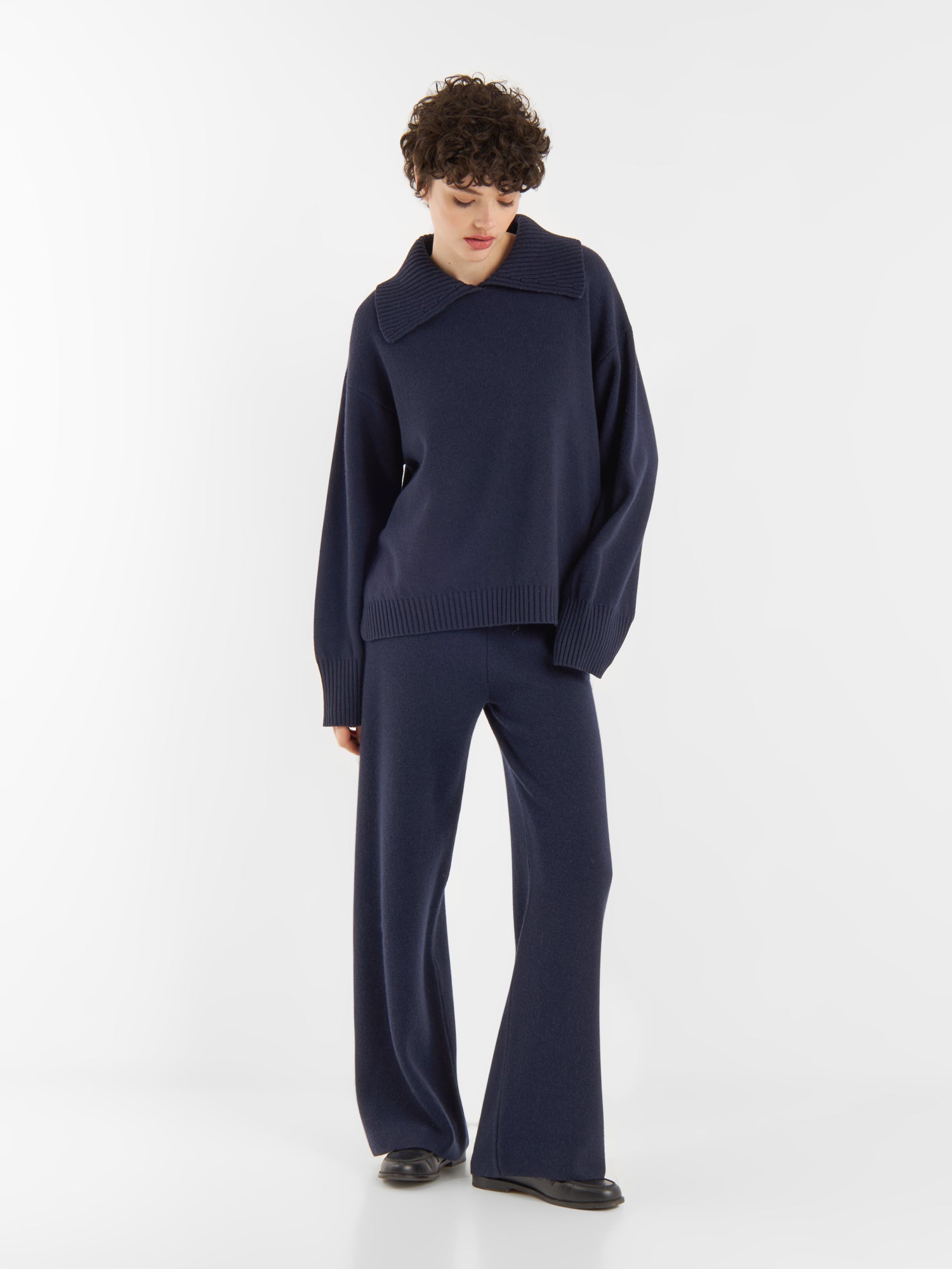 Eine Person mit kurzen lockigen Haaren trägt einen locker sitzenden Pullover Polo Merino dunkelblau mit breitem Kragen und eine dazu passende marineblaue Hose mit weitem Bein. Sie steht vor einem schlichten weißen Hintergrund und schaut nach unten.