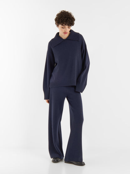 Eine Person mit kurzen lockigen Haaren trägt einen locker sitzenden Pullover Polo Merino dunkelblau mit breitem Kragen und eine dazu passende marineblaue Hose mit weitem Bein. Sie steht vor einem schlichten weißen Hintergrund und schaut nach unten.