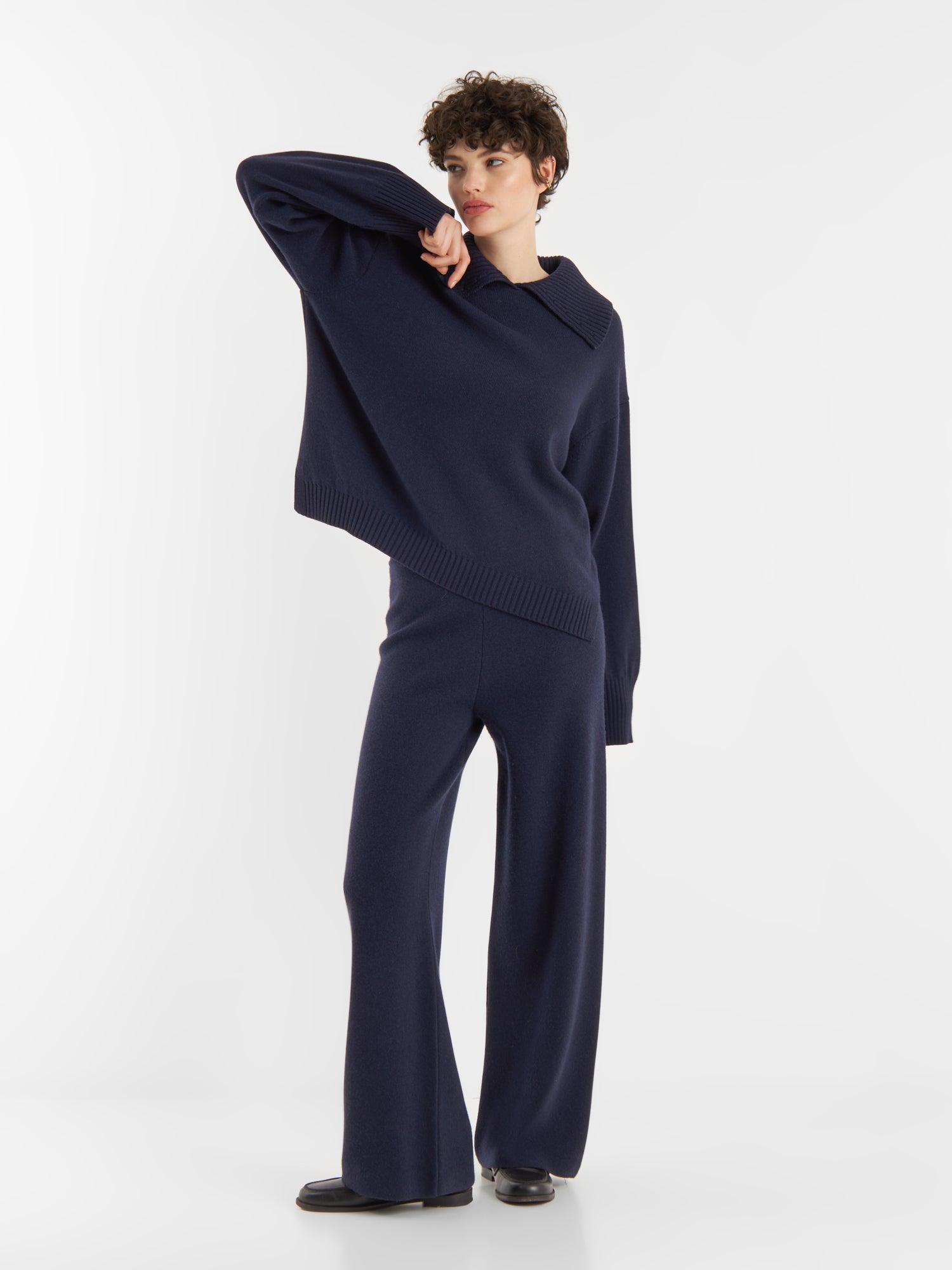 Eine Person posiert vor einem weißen Hintergrund und trägt einen locker sitzenden Pullover Polo Merino dunkelblau mit weiten Ärmeln, eine passende Hose mit weitem Bein und schwarze Schuhe.