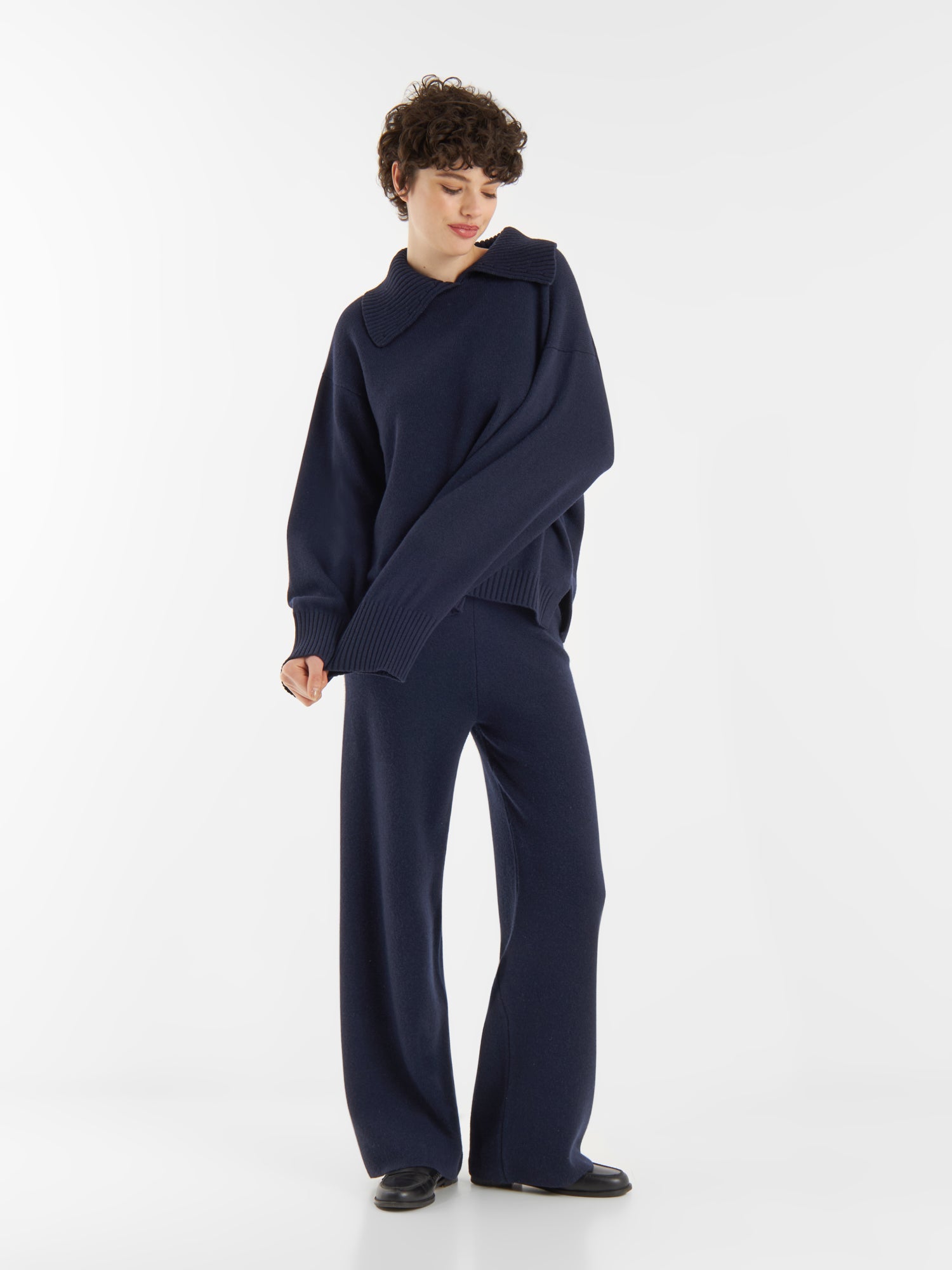 Eine Person steht vor einem weißen Hintergrund und trägt einen locker sitzenden Pullover Polo Merino dunkelblau mit extralangen Ärmeln und eine passende Hose mit weitem Bein, gepaart mit schwarzen Schuhen.
