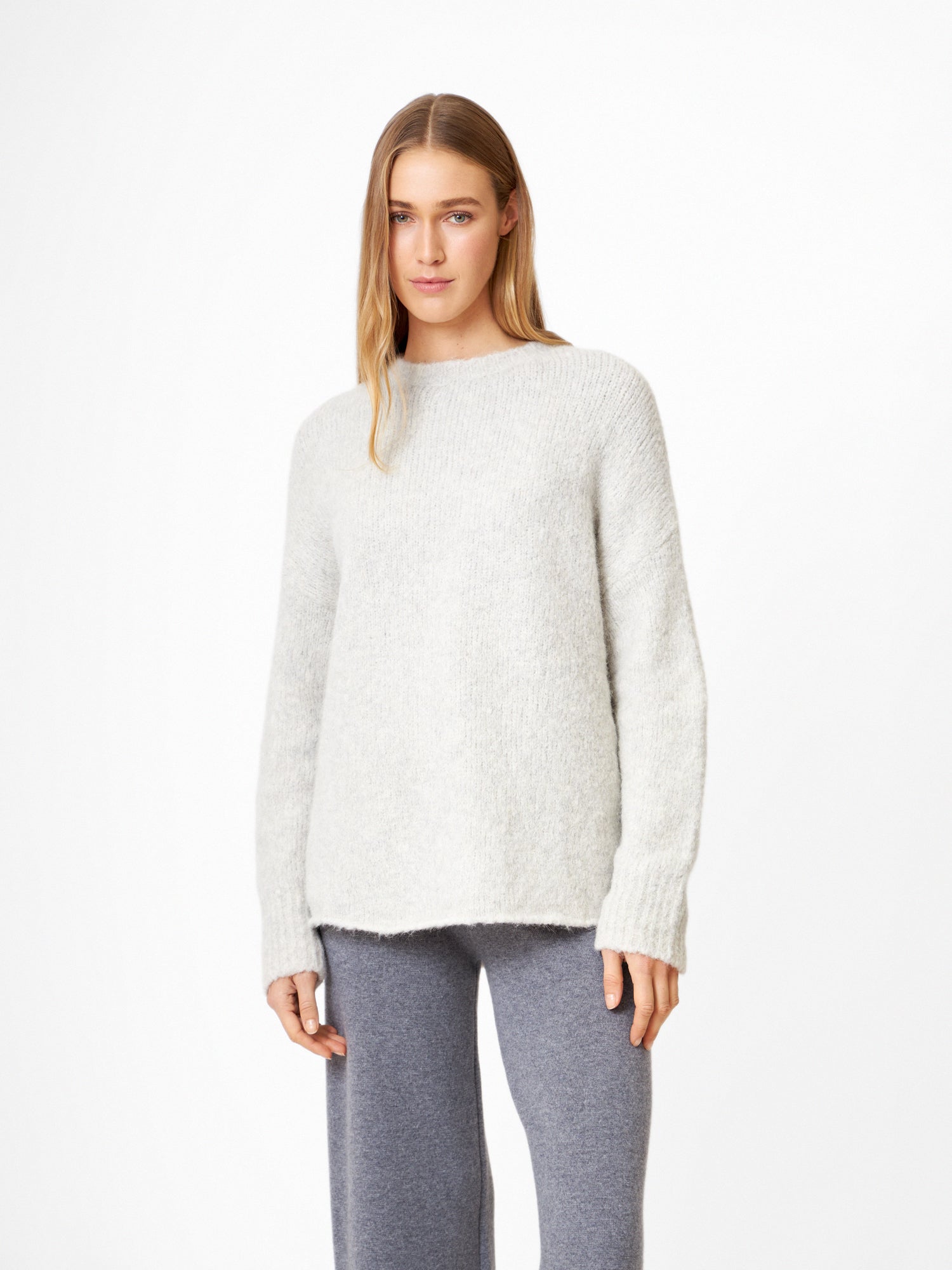 Eine Frau mit glattem, hellbraunem Haar und neutralem Gesichtsausdruck steht vor einem weißen Hintergrund und trägt den Pullover LIV in Hellgrau und eine locker sitzende, dunkelgraue Hose.