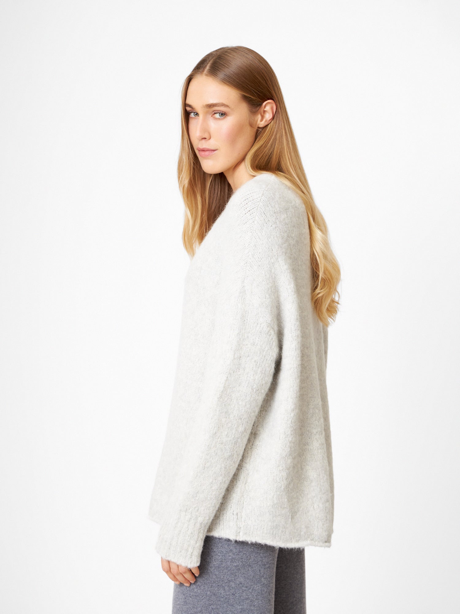 Eine Frau mit langen blonden Haaren trägt den Pullover LIV in Hellgrau und eine dazu passende graue Hose. Sie steht seitlich und schaut in die Kamera vor einem schlichten weißen Hintergrund.