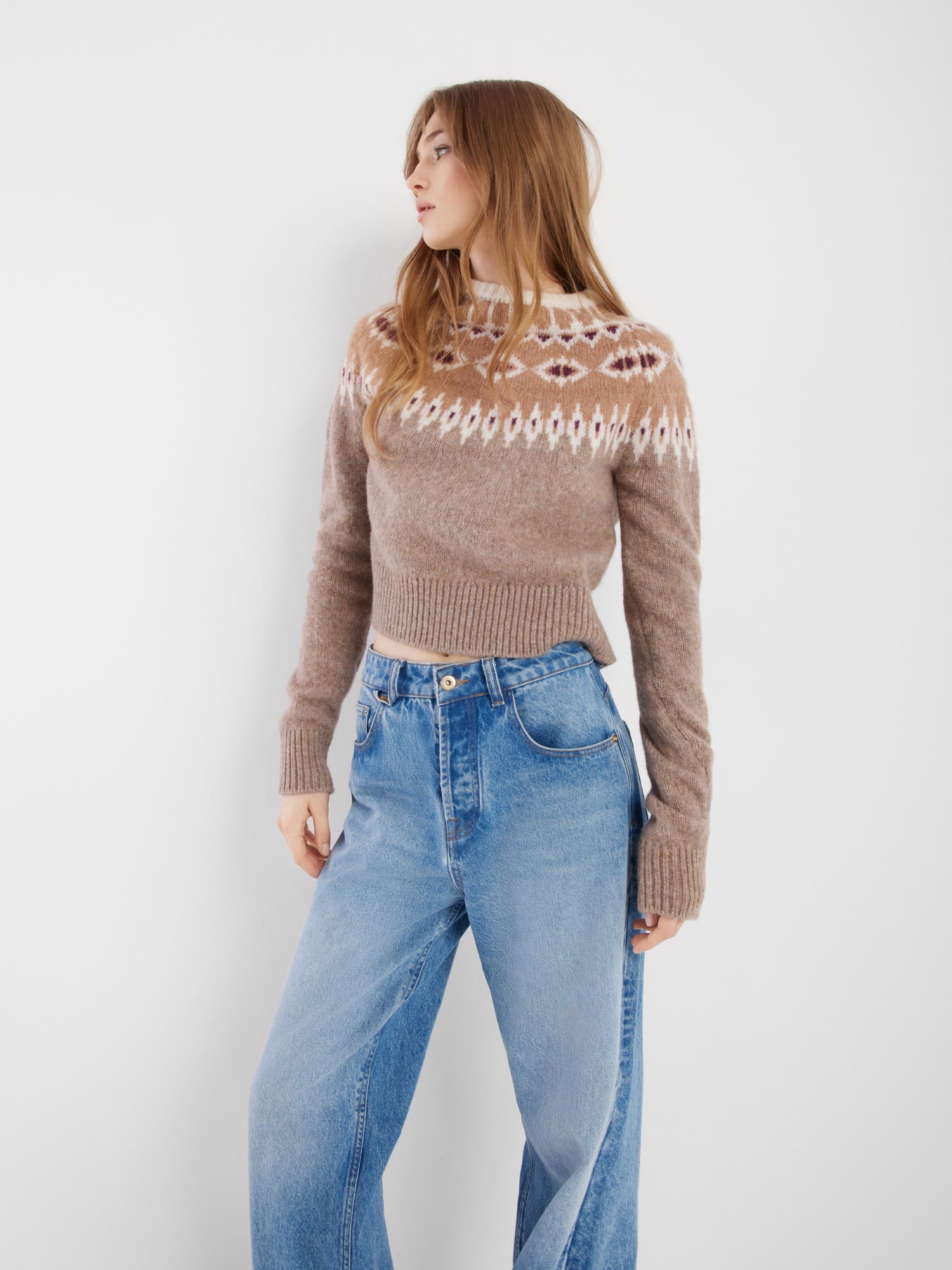 Eine Frau mit langen braunen Haaren trägt den Pullover Norweger Vol.2 Beige Burgundy und eine Jeans mit hoher Taille. Sie steht vor einem schlichten weißen Hintergrund und blickt zur Seite.