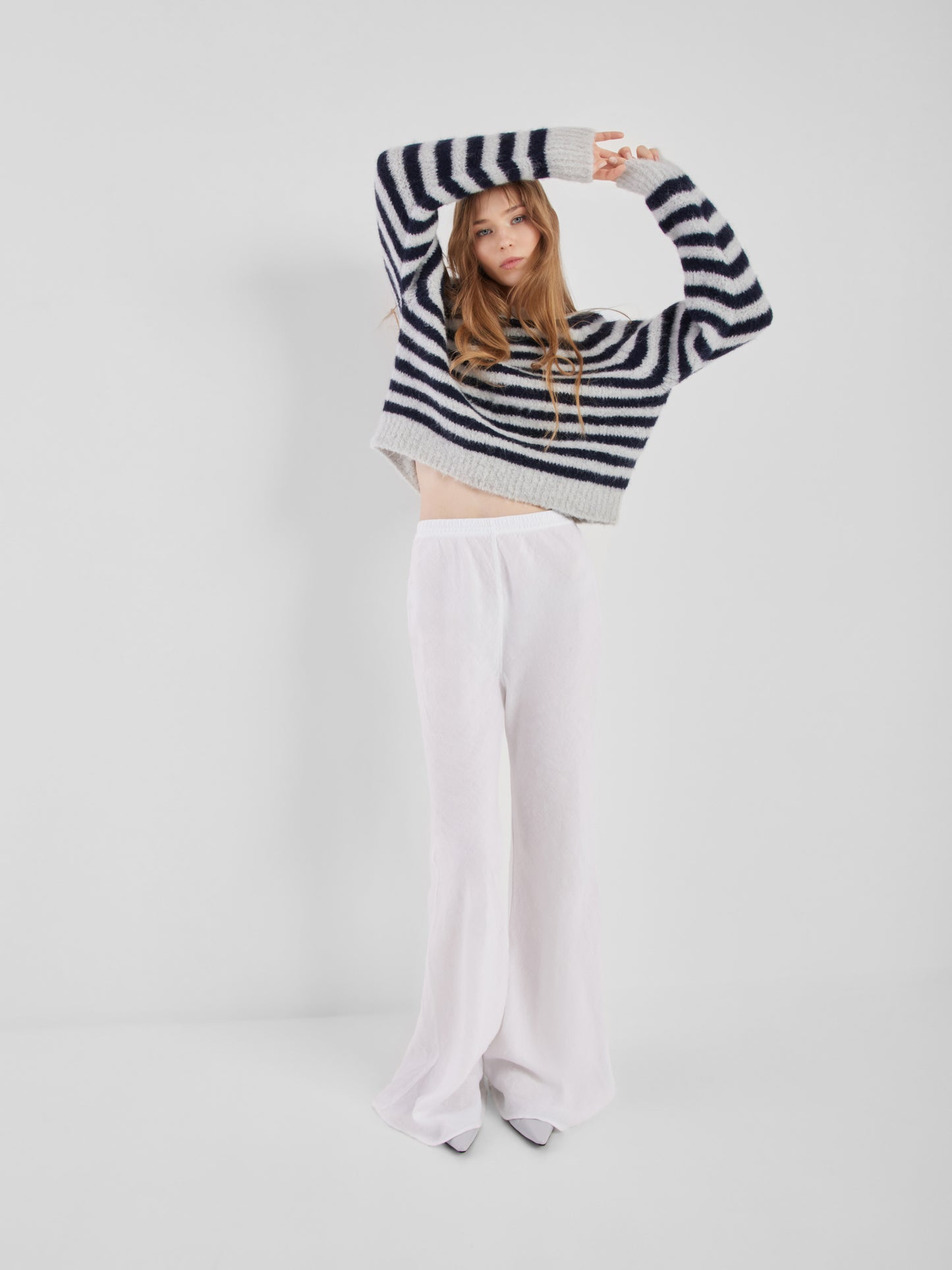 Eine Frau mit langen Haaren posiert vor einem unifarbenen Hintergrund. Sie trägt den Pullover Blue Stripe mit langen Ärmeln und eine weite, hochgeschlossene weiße Hose. Sie hebt die Arme über den Kopf und blickt in die Kamera.