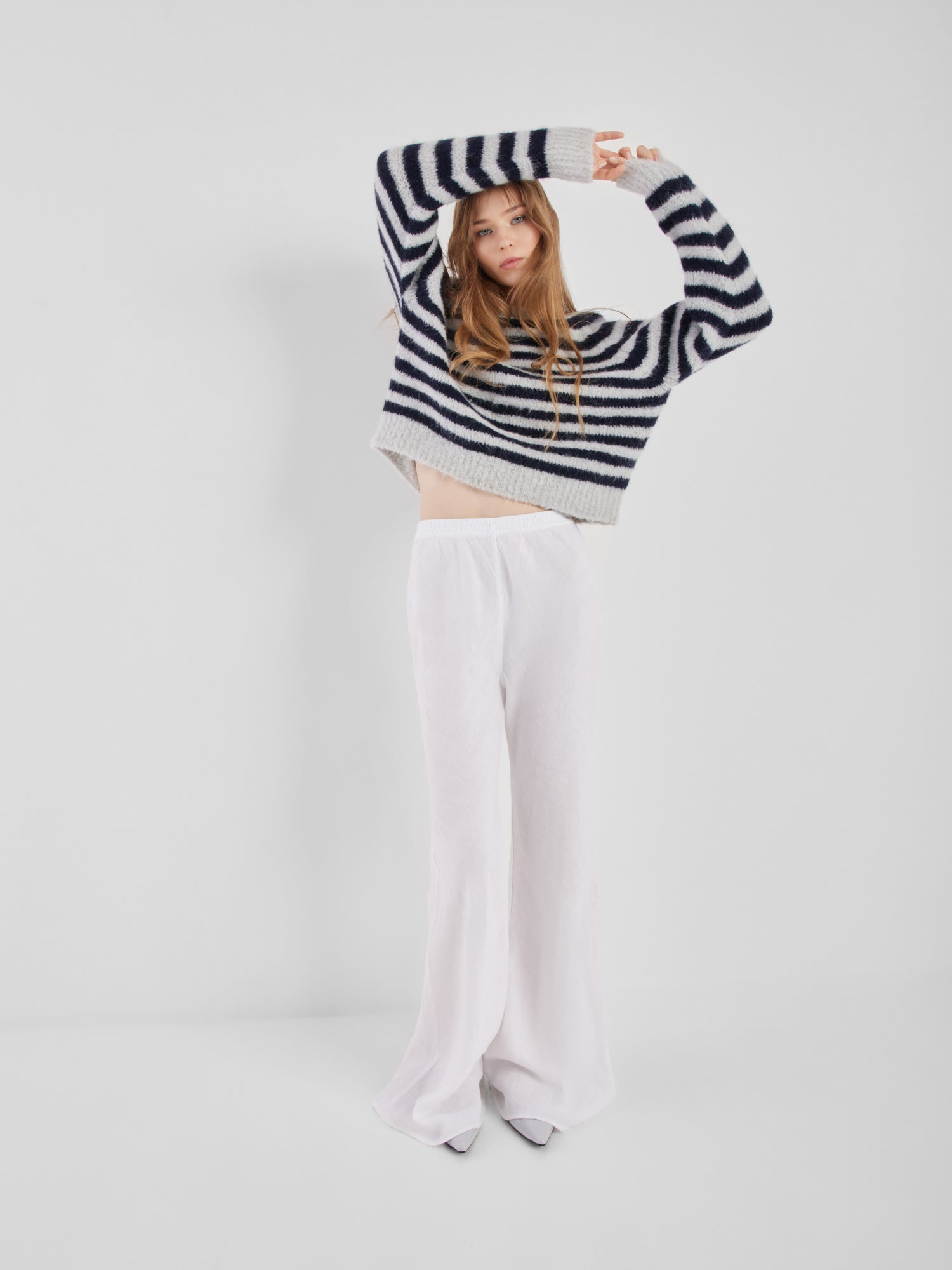 Eine Frau mit langen Haaren posiert vor einem unifarbenen Hintergrund. Sie trägt den Pullover Blue Stripe mit langen Ärmeln und eine weite, hochgeschlossene weiße Hose. Sie hebt die Arme über den Kopf und blickt in die Kamera.
