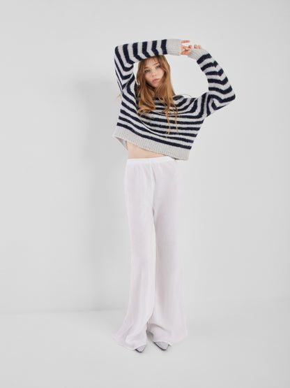 Eine Frau mit langen Haaren posiert vor einem unifarbenen Hintergrund. Sie trägt den Pullover Blue Stripe mit langen Ärmeln und eine weite, hochgeschlossene weiße Hose. Sie hebt die Arme über den Kopf und blickt in die Kamera.