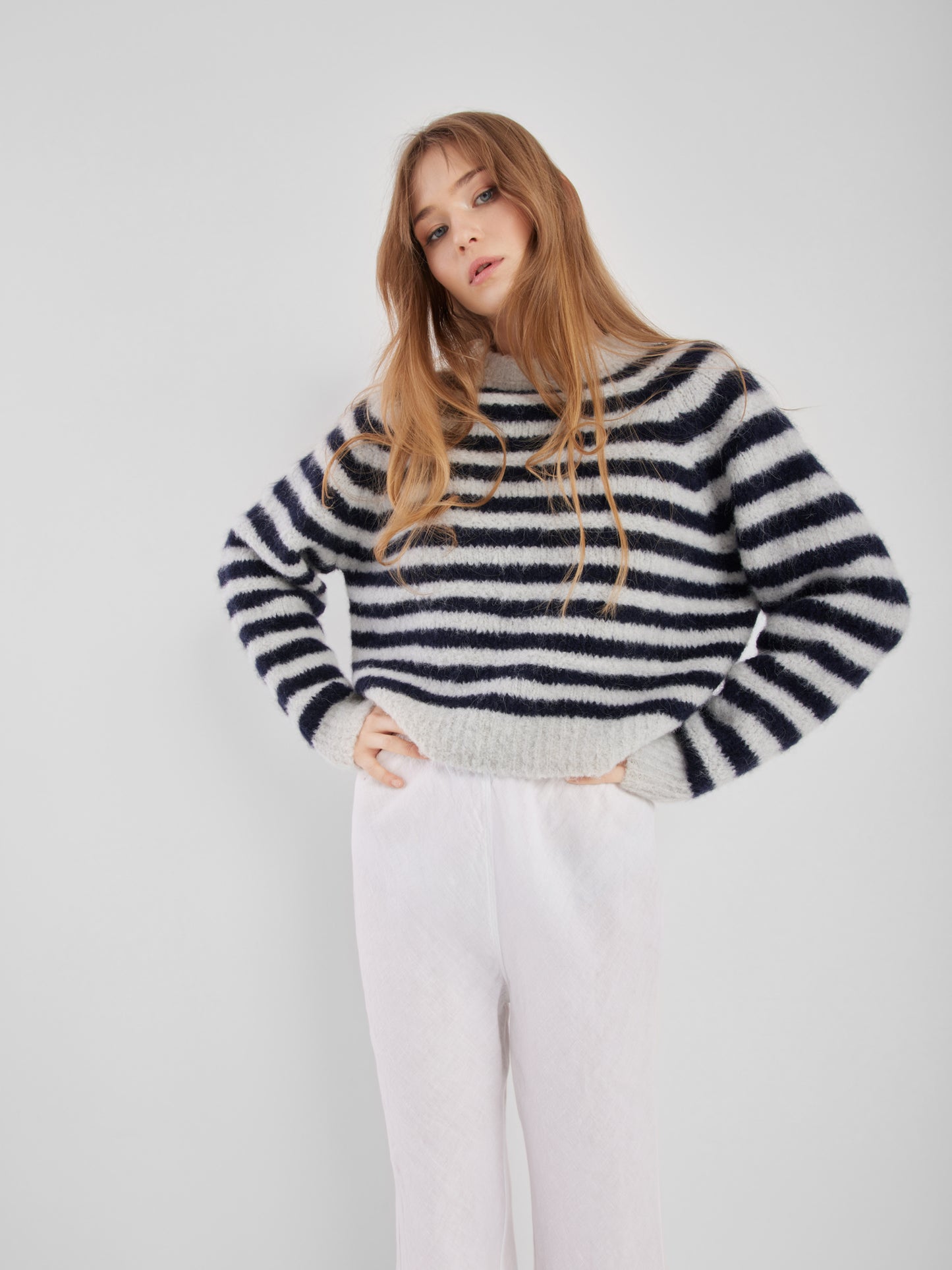 Eine Person mit langem, hellbraunem Haar steht vor einem schlichten, hellen Hintergrund. Sie trägt einen blau gestreiften Pullover und eine weiße Hose, die Hände in die Hüften gestützt.