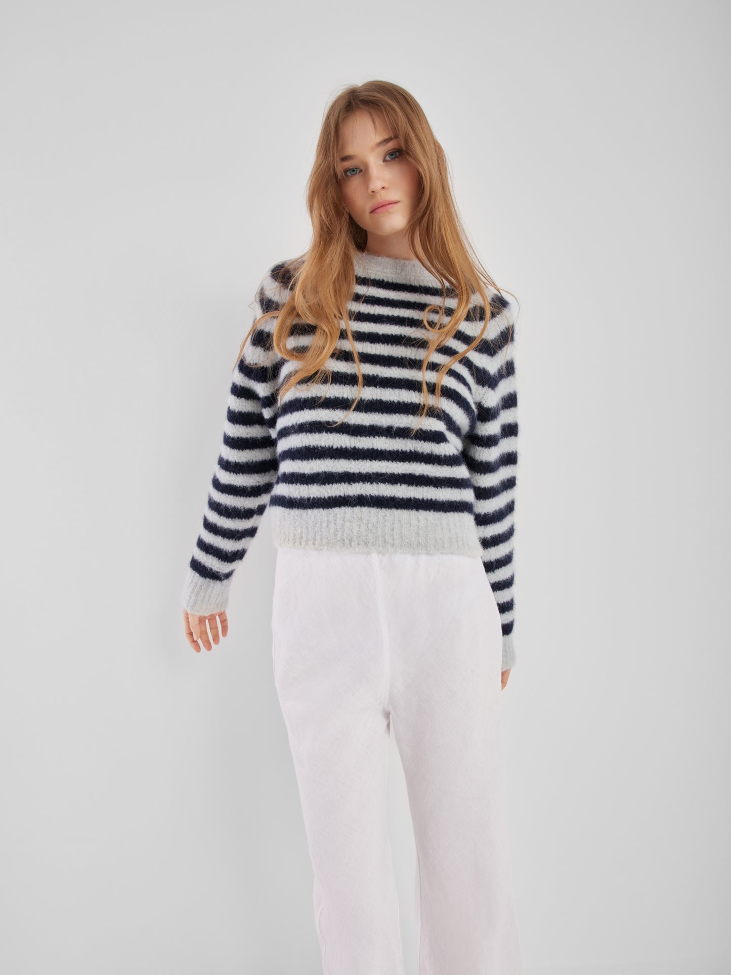 Eine Frau mit langen, hellbraunen Haaren trägt den Pullover Blue Stripe und eine weiße Hose vor einem schlichten weißen Hintergrund.