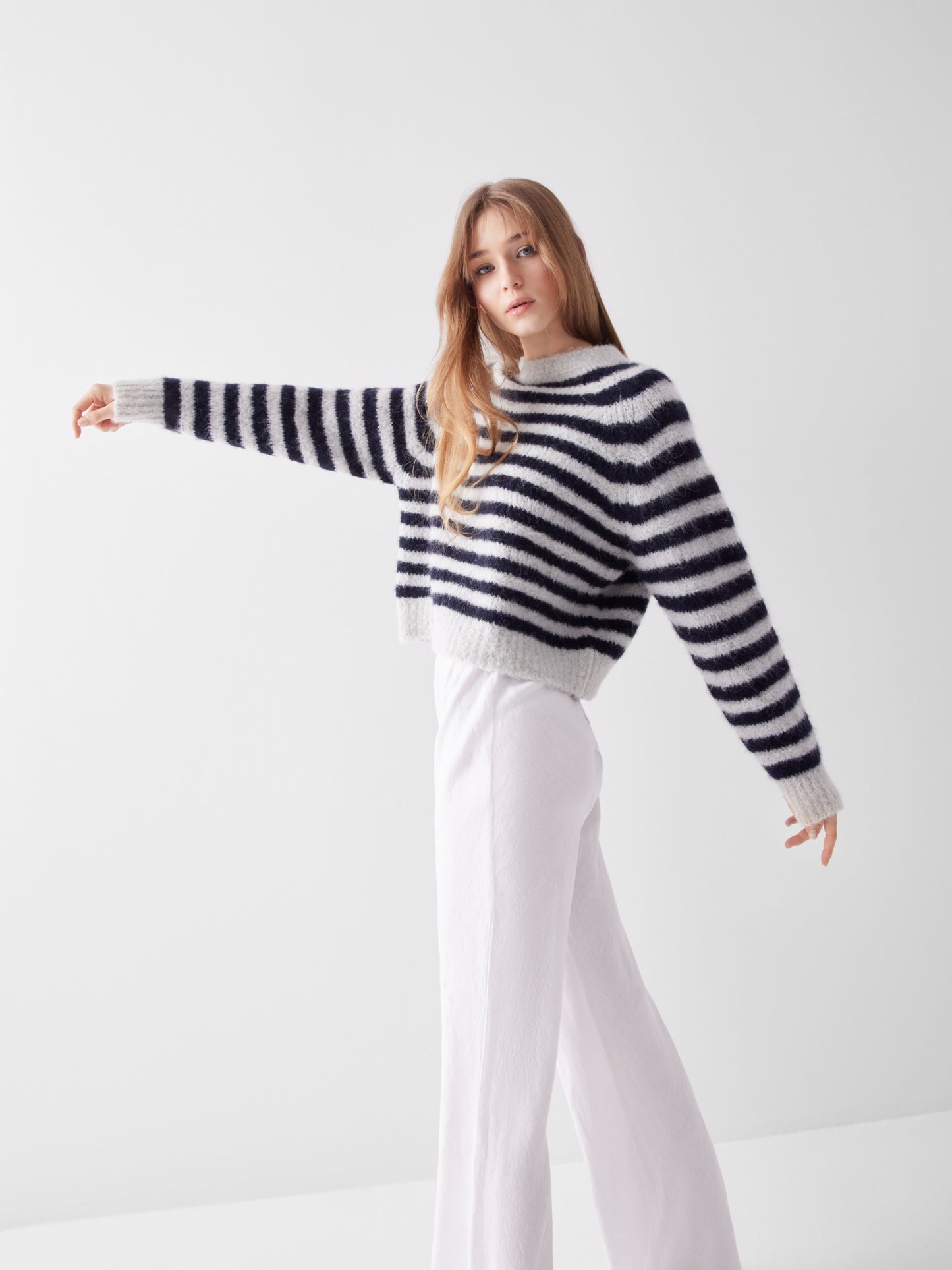 Eine Person mit langen Haaren trägt den Pullover Blue Stripe und eine weiße Hose mit weitem Bein. Sie steht vor einem einfarbig weißen Hintergrund und streckt einen Arm zur Seite.