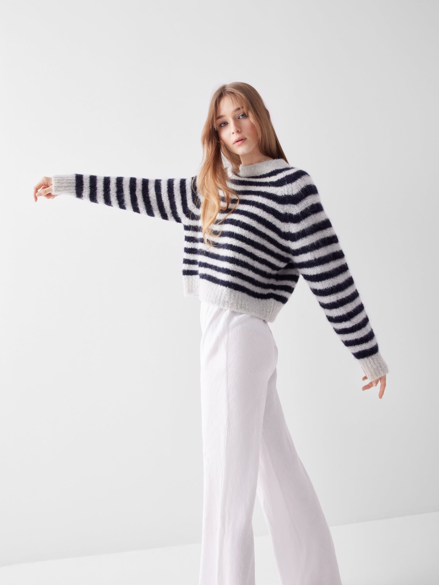 Eine Person mit langen Haaren trägt den Pullover Blue Stripe und eine weiße Hose mit weitem Bein. Sie steht vor einem einfarbig weißen Hintergrund und streckt einen Arm zur Seite.