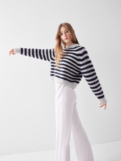 Eine Person mit langen Haaren trägt den Pullover Blue Stripe und eine weiße Hose mit weitem Bein. Sie steht vor einem einfarbig weißen Hintergrund und streckt einen Arm zur Seite.