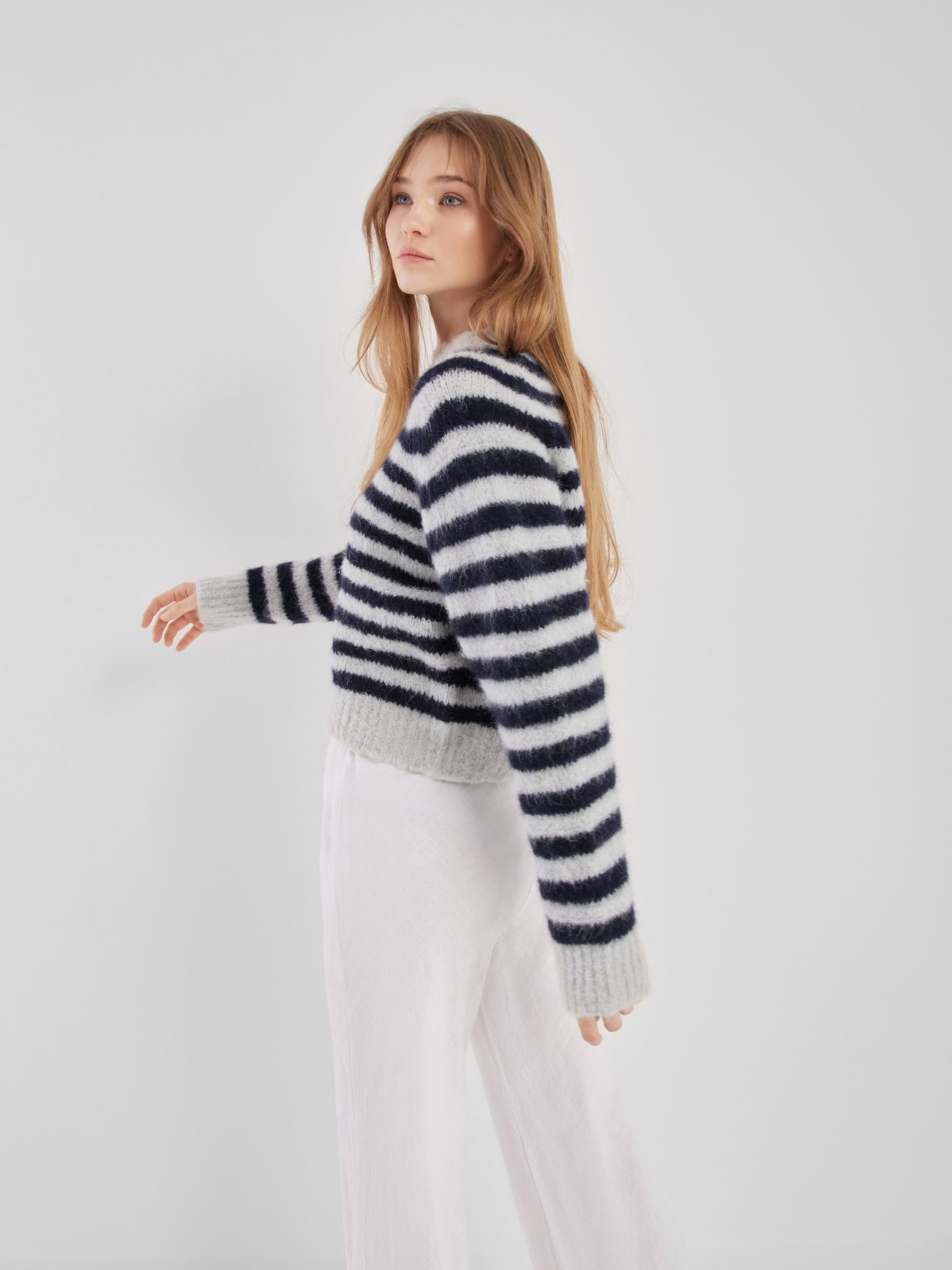 Eine Frau mit langen hellbraunen Haaren steht seitlich vor einem schlichten weißen Hintergrund. Sie trägt den Pullover Blue Stripe und eine weite weiße Hose.