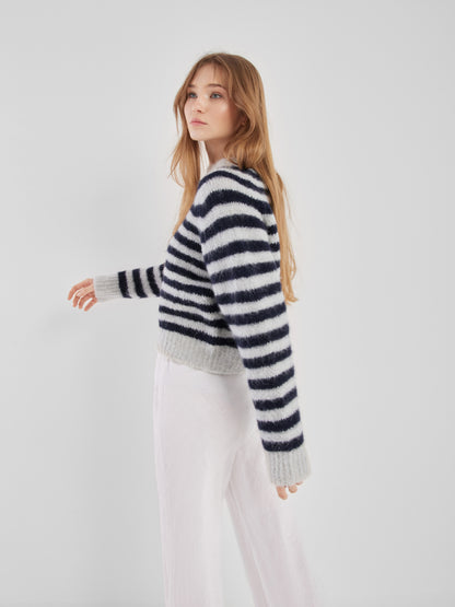 Eine Frau mit langen hellbraunen Haaren steht seitlich vor einem schlichten weißen Hintergrund. Sie trägt den Pullover Blue Stripe und eine weite weiße Hose.