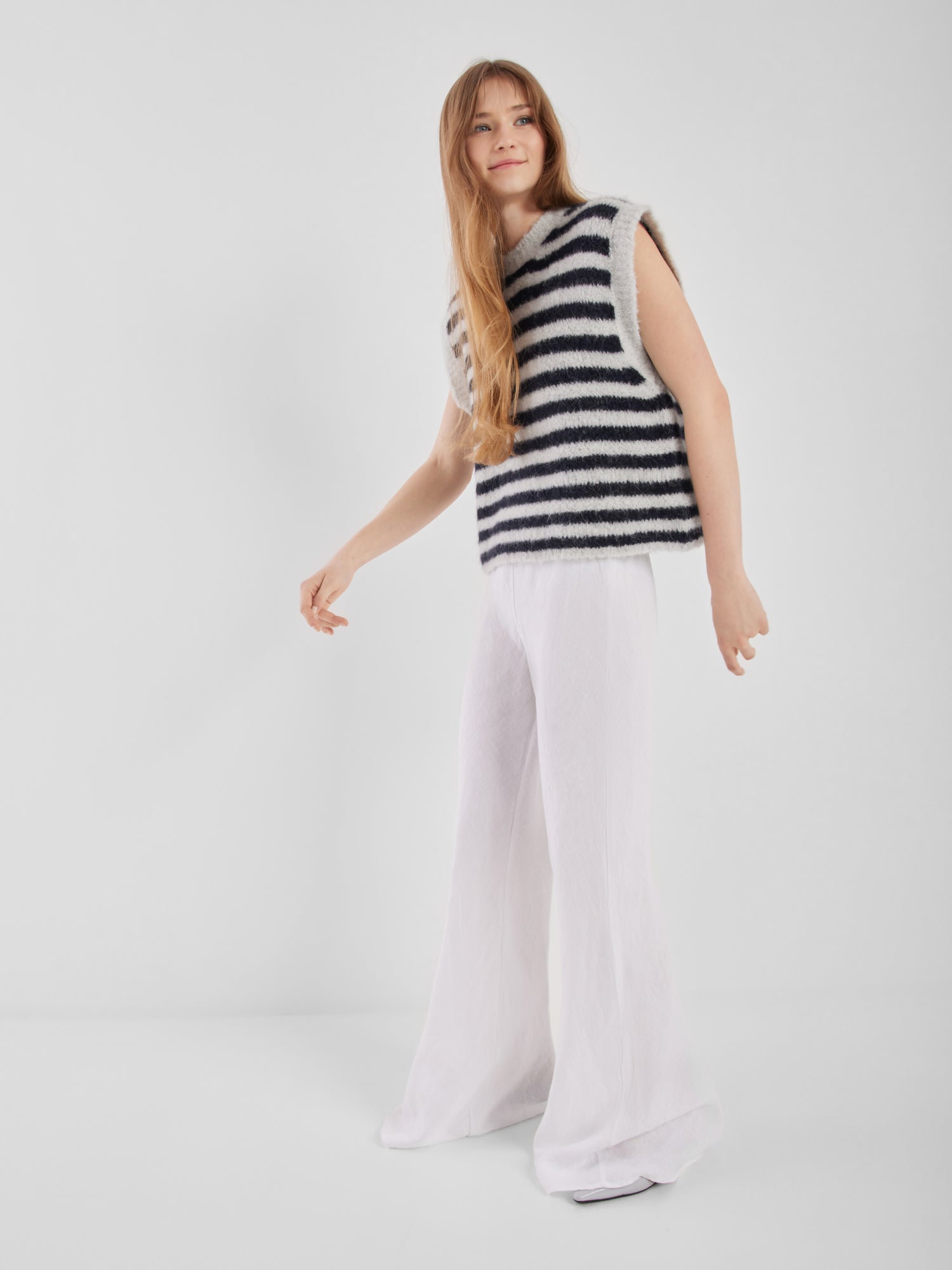 Eine Frau mit langen Haaren steht vor einem schlichten weißen Hintergrund, lächelt sanft und blickt zur Seite. Sie trägt den Pullunder Blue Stripe und eine weit geschnittene weiße Hose.