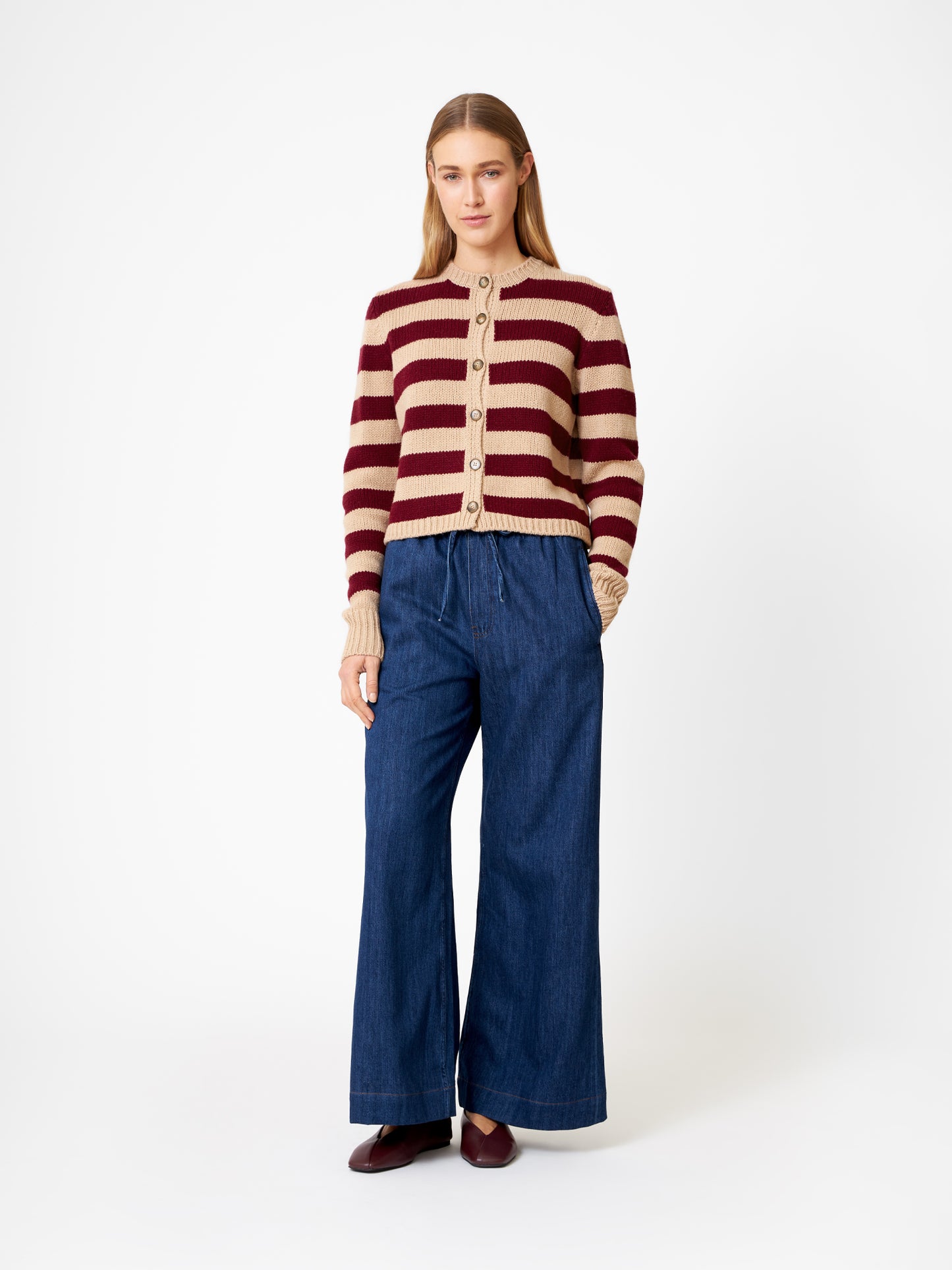 Eine Frau mit langem, glattem Haar und neutralem Gesichtsausdruck steht vor einem weißen Hintergrund und trägt die Strickjacke Jacket Stripe Burgundy Stripe (Made in Italy), weite Blue Jeans und braune Schuhe.