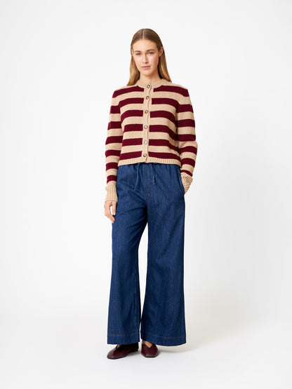 Eine Frau mit langem, glattem Haar und neutralem Gesichtsausdruck steht vor einem weißen Hintergrund und trägt die Strickjacke Jacket Stripe Burgundy Stripe (Made in Italy), weite Blue Jeans und braune Schuhe.
