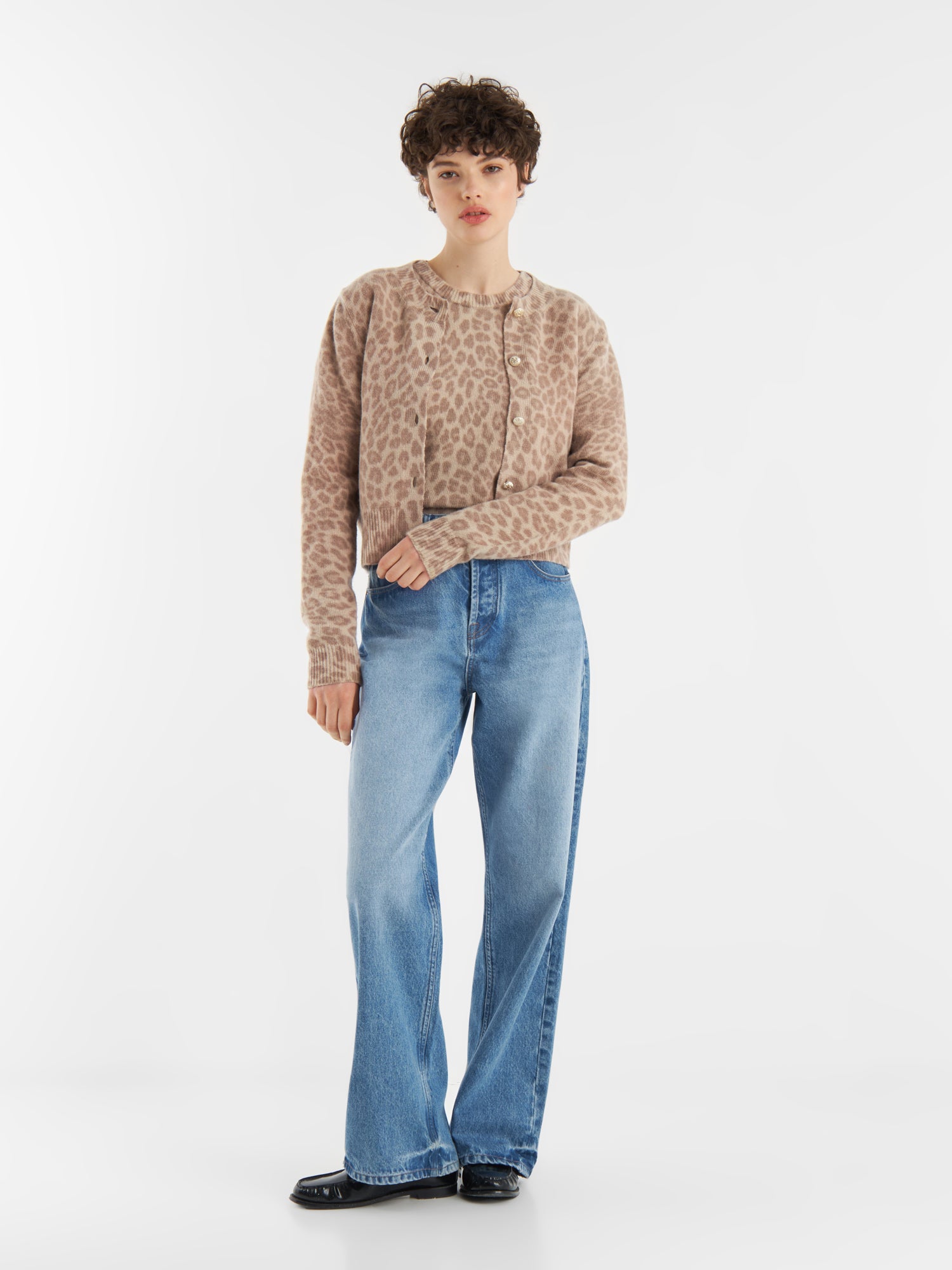 Eine Person mit kurzen lockigen Haaren trägt eine beigefarbene Strickjacke mit Leo-Print aus weicher Merinowolle, gepaart mit hellblauen Jeans mit weitem Bein und schwarzen Schuhen, vor einem schlichten weißen Hintergrund.