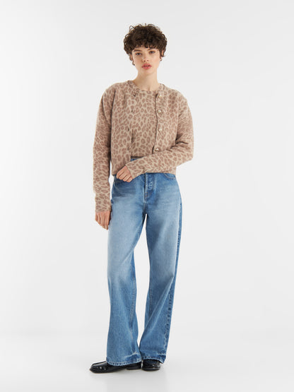 Eine Person mit kurzen lockigen Haaren trägt eine beigefarbene Strickjacke mit Leo-Print aus weicher Merinowolle, gepaart mit hellblauen Jeans mit weitem Bein und schwarzen Schuhen, vor einem schlichten weißen Hintergrund.